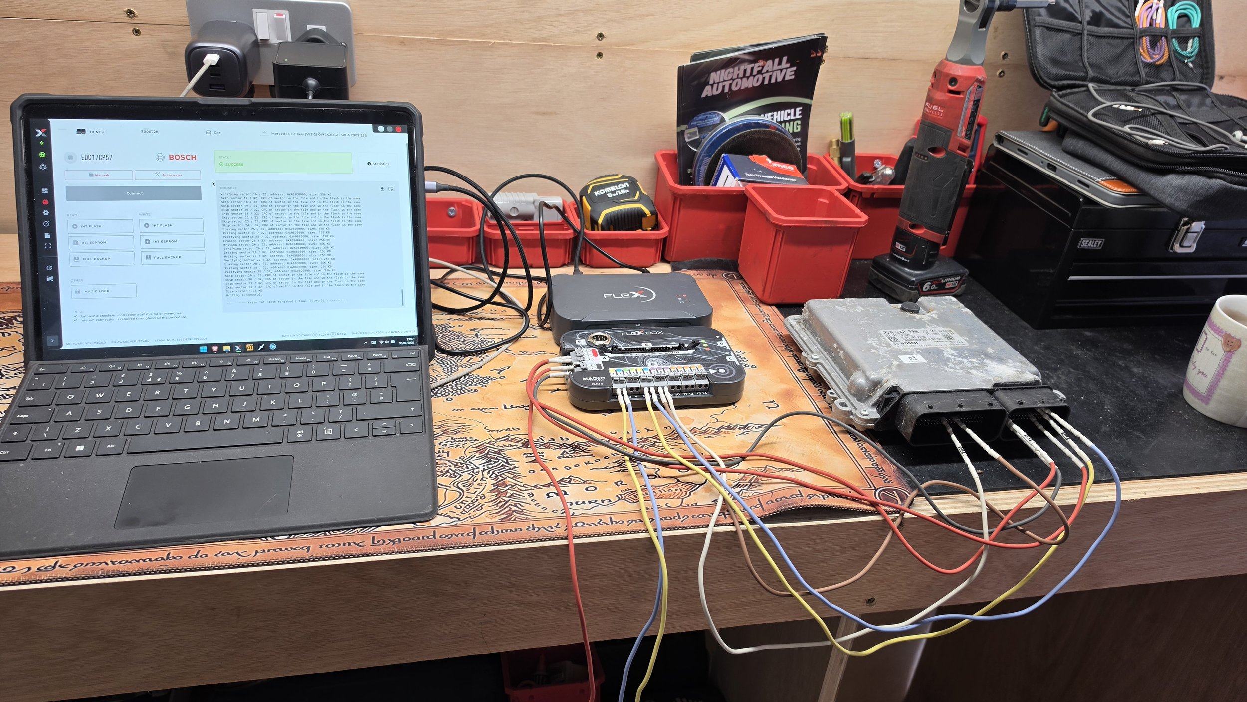 A workbench with a tablet, electronic testing equipment, wires, and automotive repair tools including a drill, a laptop, a circuit board, and a can of soda, set against a wooden wall. tuned by Nightfall Automotive