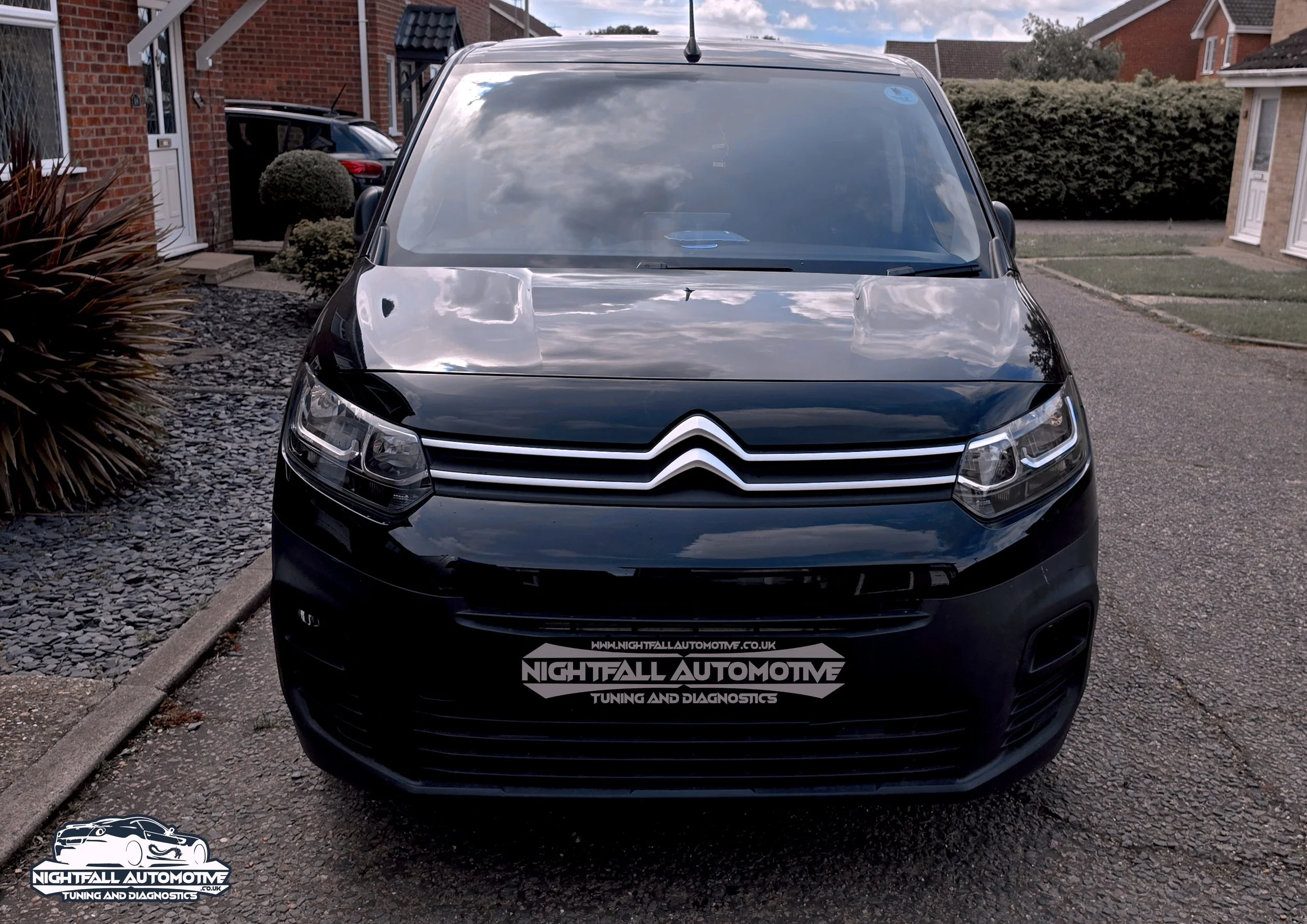 Black car parked in a residential driveway with houses in the background. The car has a shiny exterior with reflections of clouds in the windows, and a logo on the front grille. There is a sticker on the windshield. tuned by Nightfall Automotive