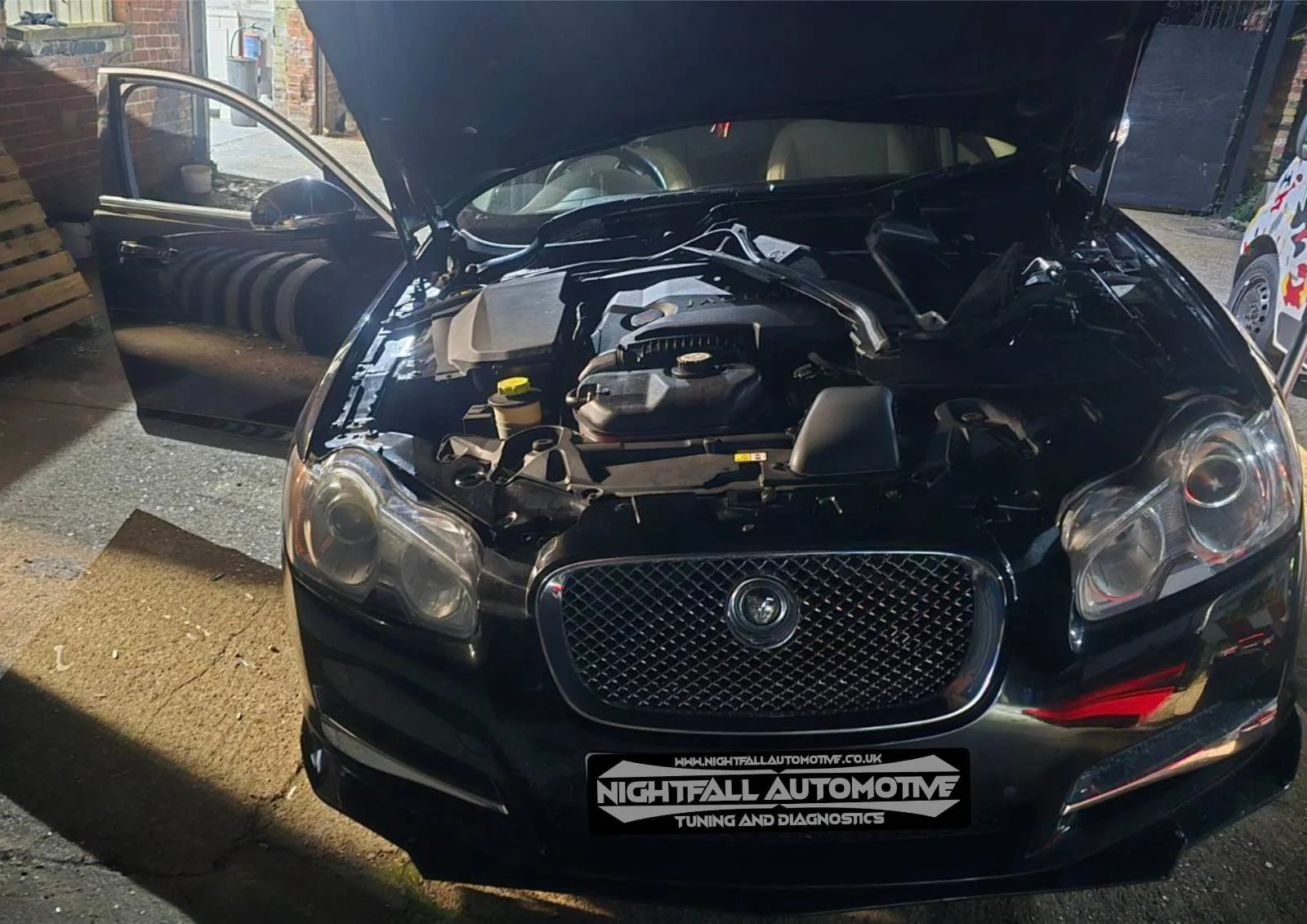 Black car with open hood parked outdoors at night, showing engine compartment, with front passenger door open and a sign on the front bumper reading 'Nightfall Automotive Tuning and Diagnostics'.