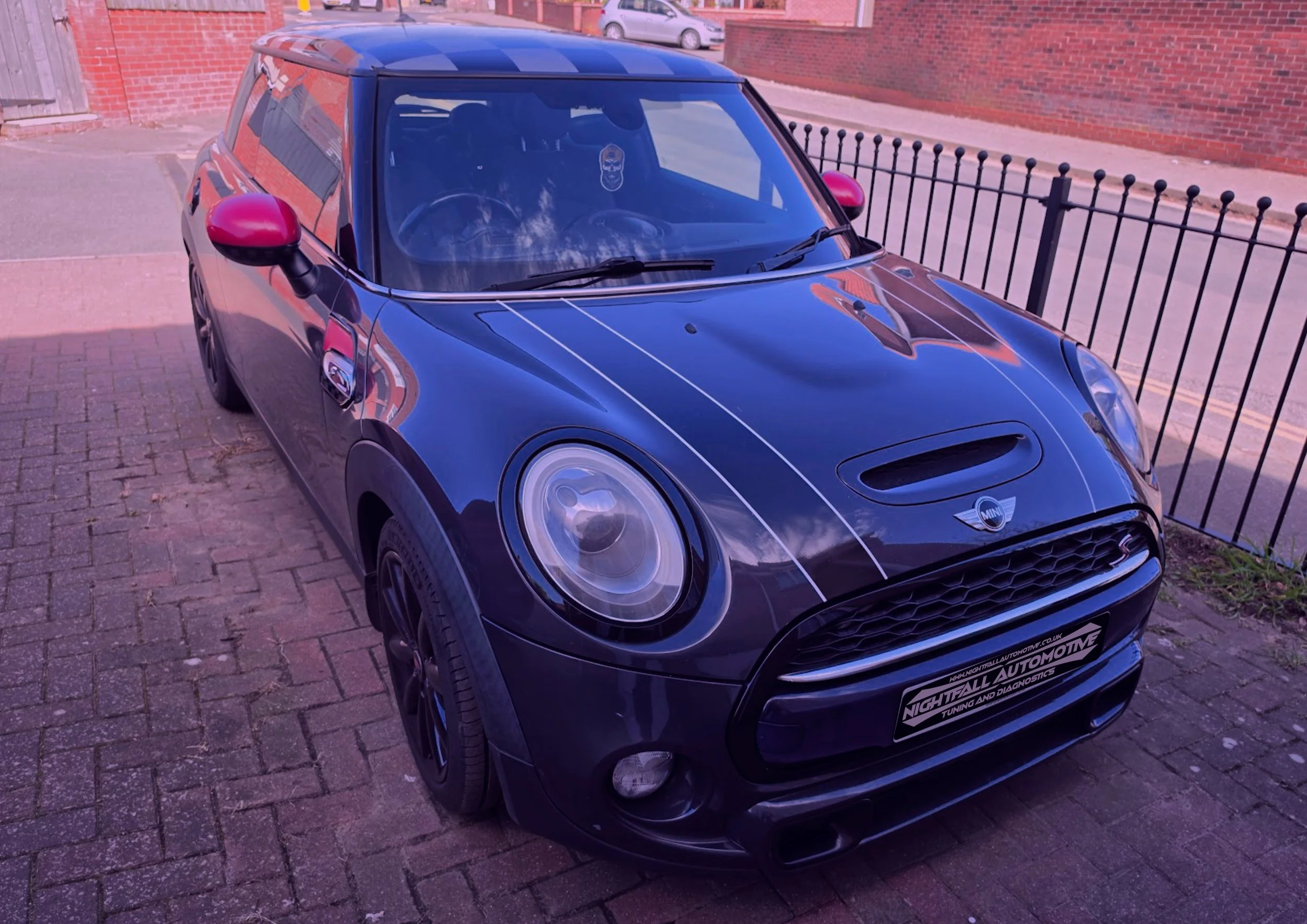 A black Mini Cooper with red side mirrors and a black hood with a vent, parked on a brick sidewalk next to a black fence and brick building. stage 1 and EGR solution by Nightfall Automotive