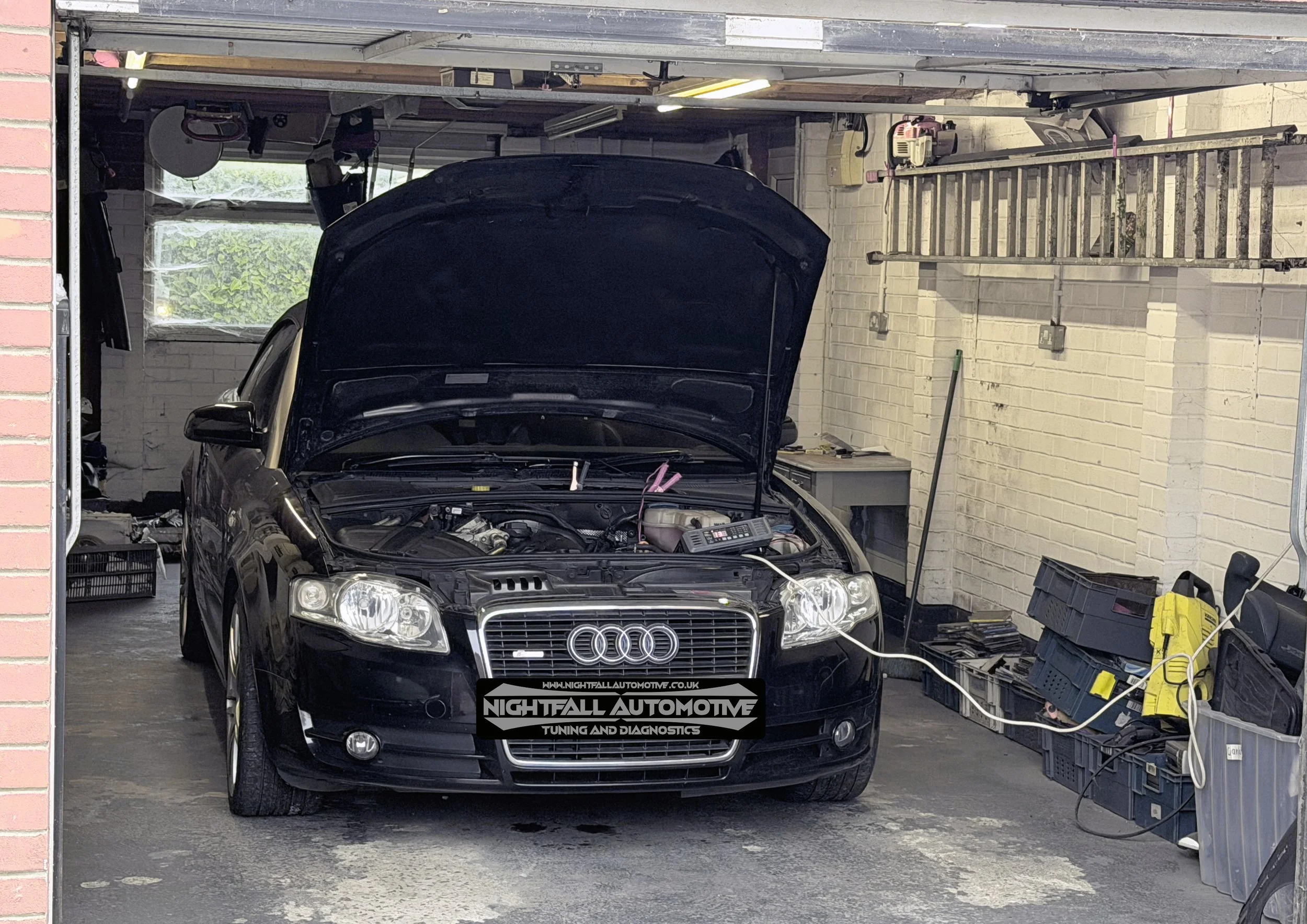 A black Audi car with its hood open inside a garage, with various tools and equipment around, and a ladder leaning against the wall. Tuned by Nightfall Automotive
