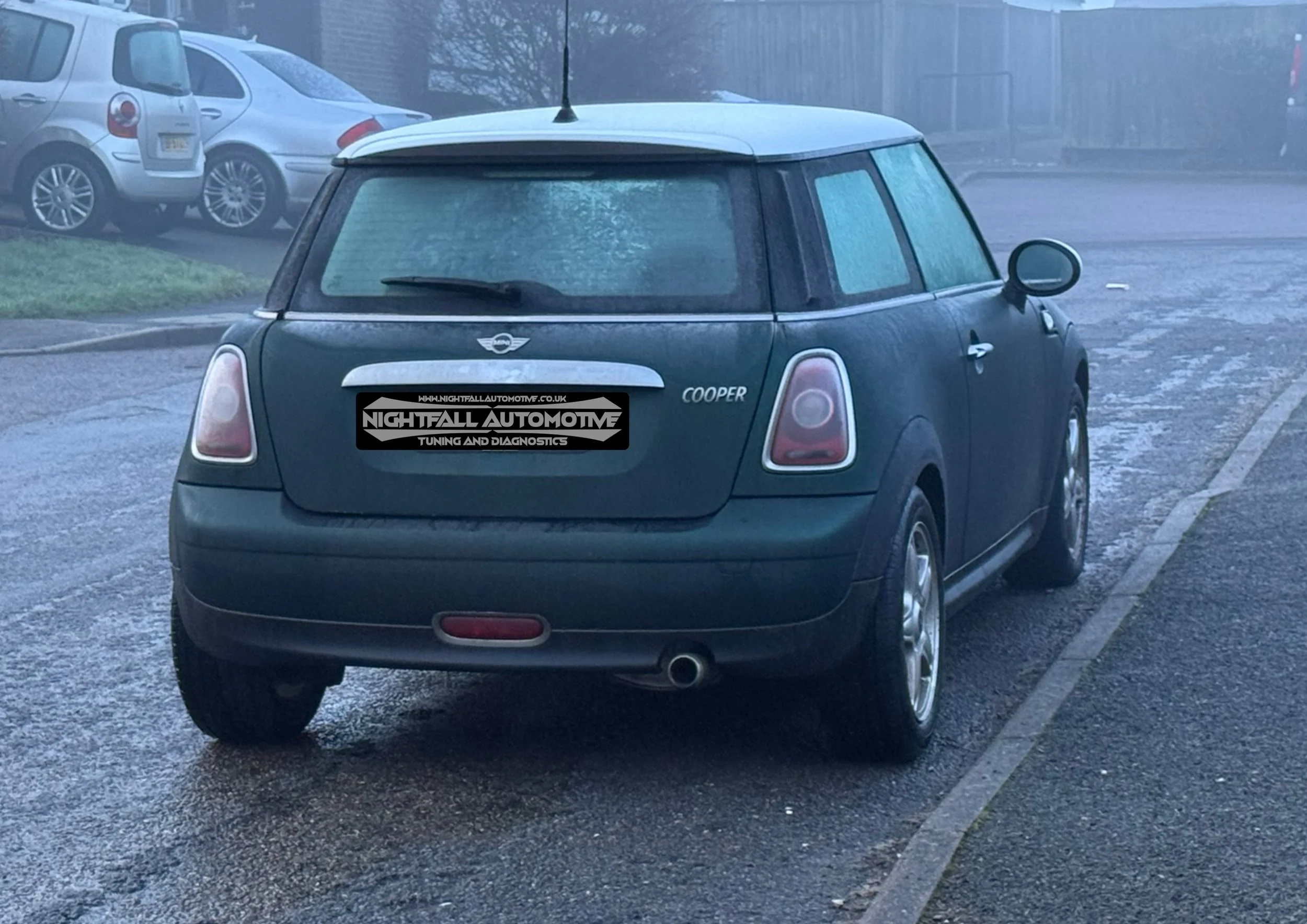 Rear view of a dark green Mini Cooper car parked on a wet street, with other cars in the background, and a sticker on the back that reads 'Nightfall Automotive Tuning and Diagnostics'.