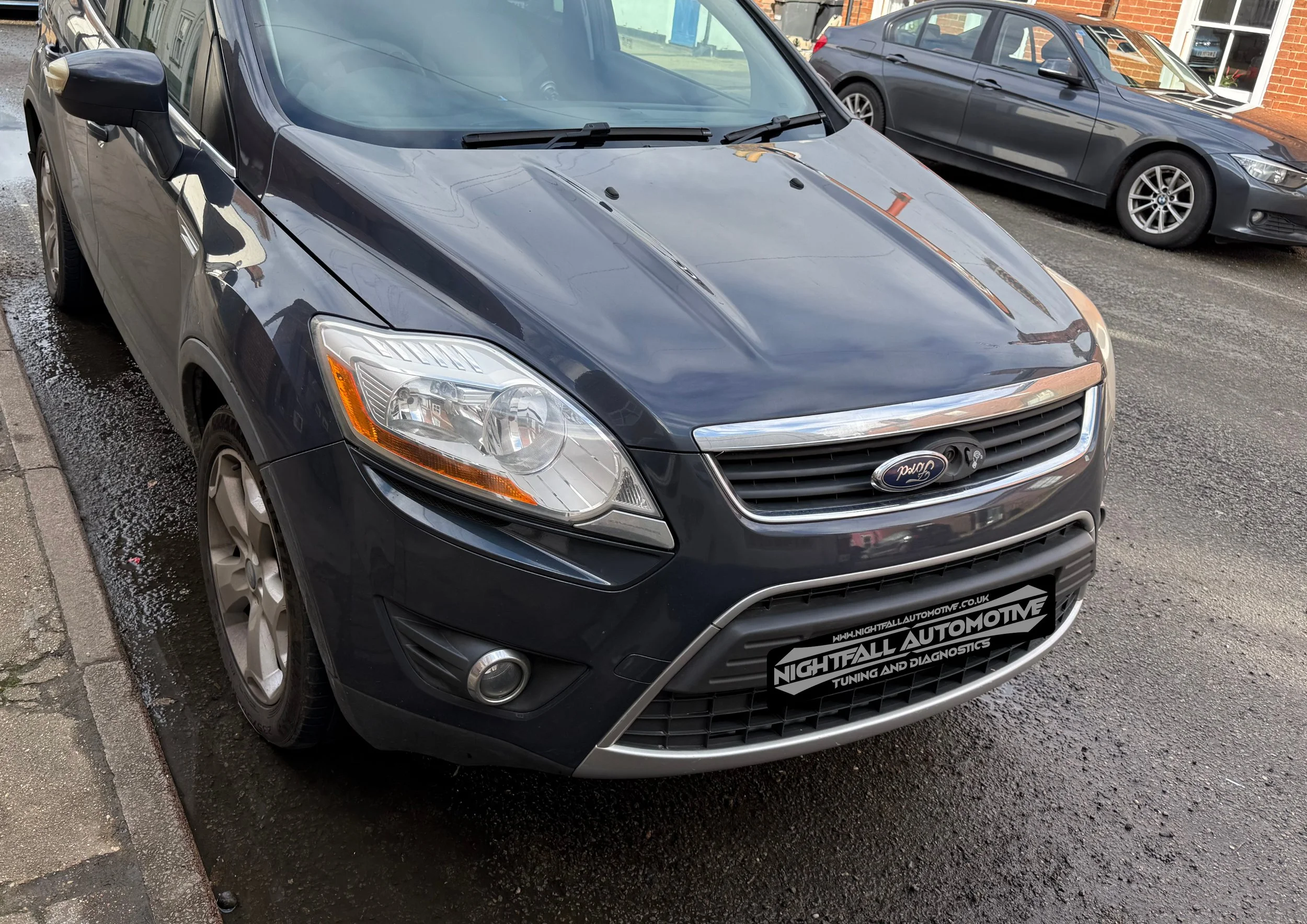 Black Ford car parked on a wet street, with other vehicles and brick buildings in the background.