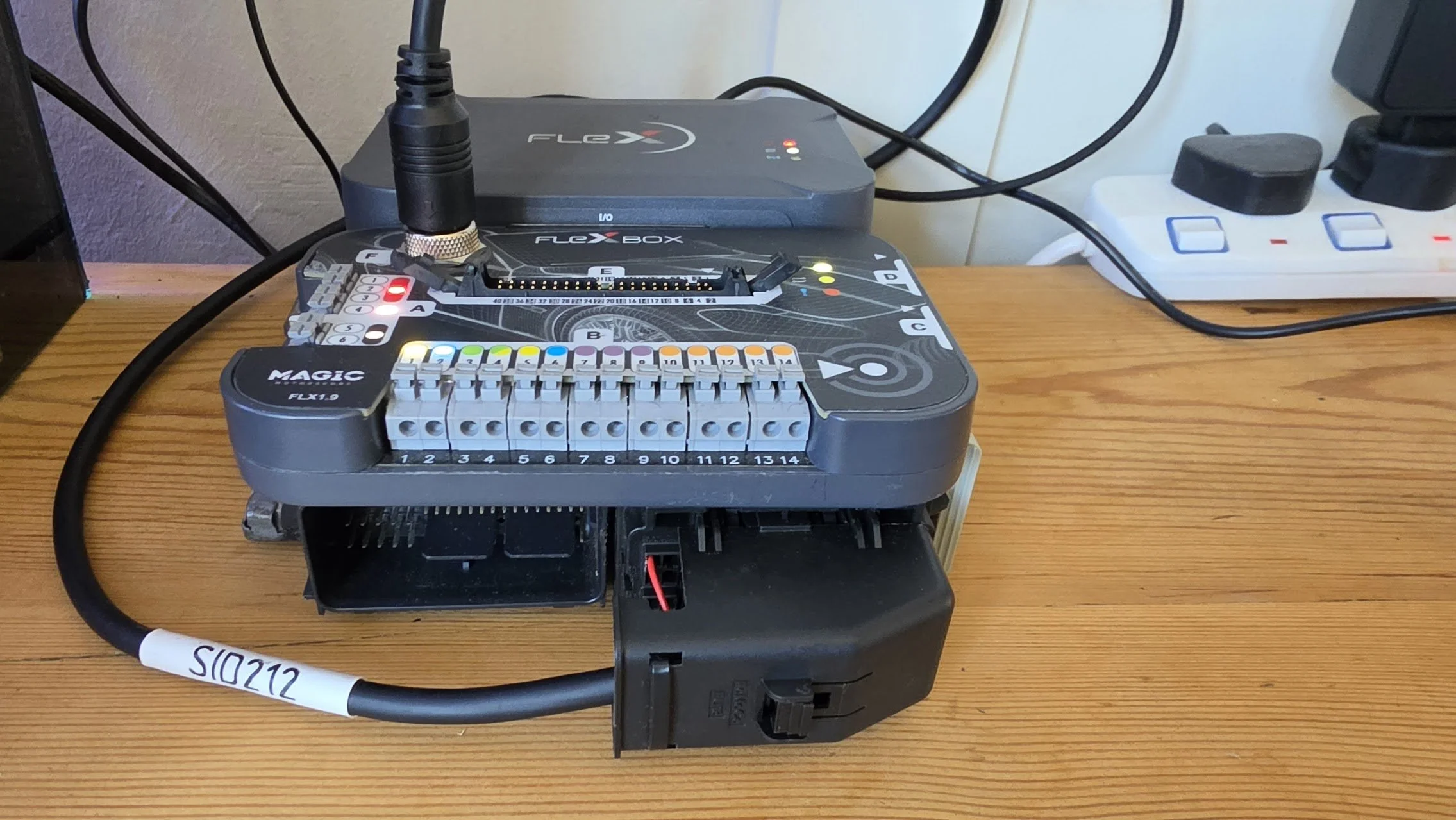 A partially assembled electronic device with wires and circuit components on a wooden desk, with a power strip in the background. Flex used by Nightfall Automotive