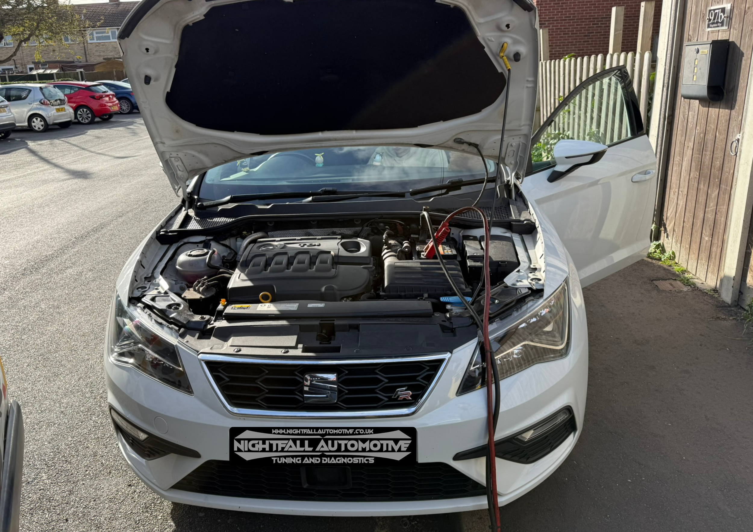 White car with open hood and battery charger connected, parked outside near a wooden fence and other vehicles.