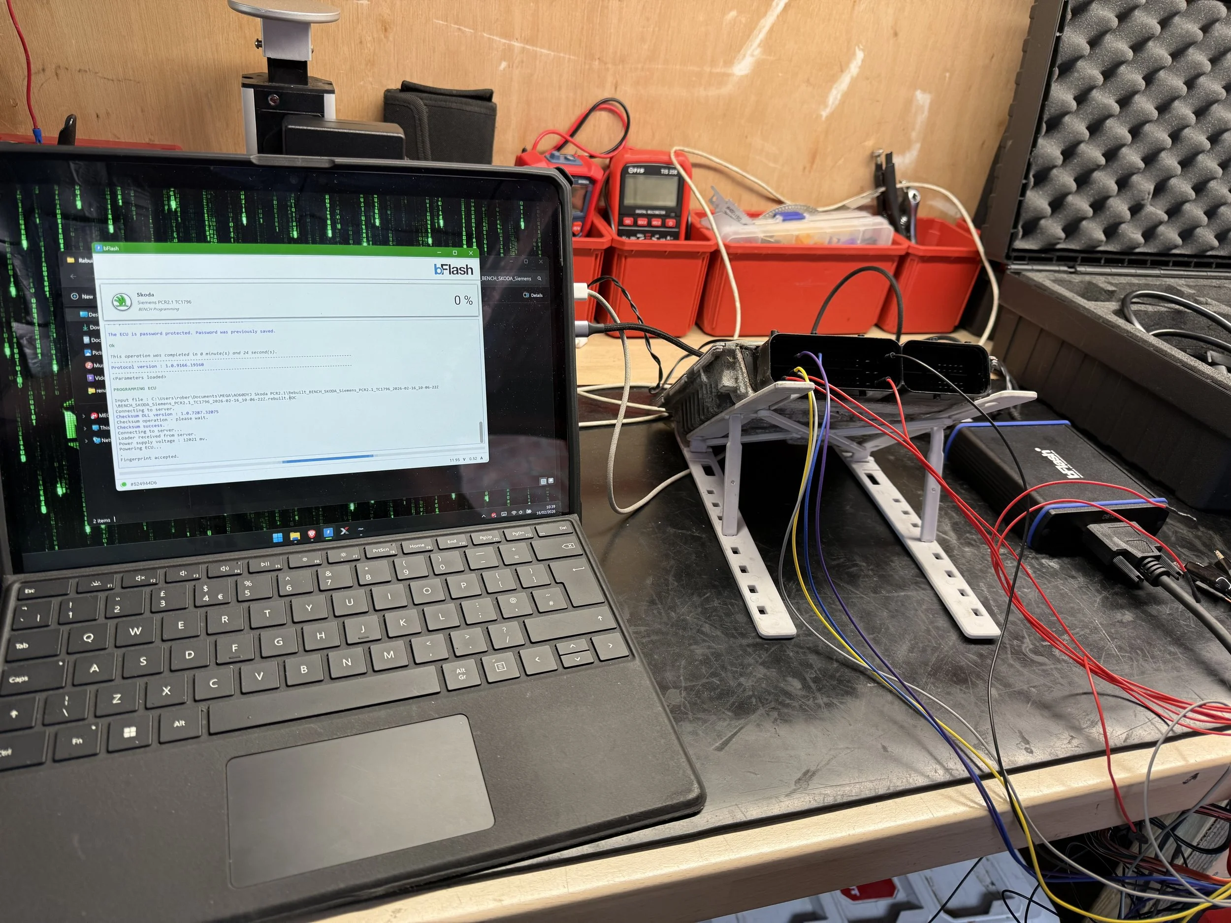 Laptop on a workbench connected to a robot with wires, surrounded by tools and equipment for electronics or robotics projects.