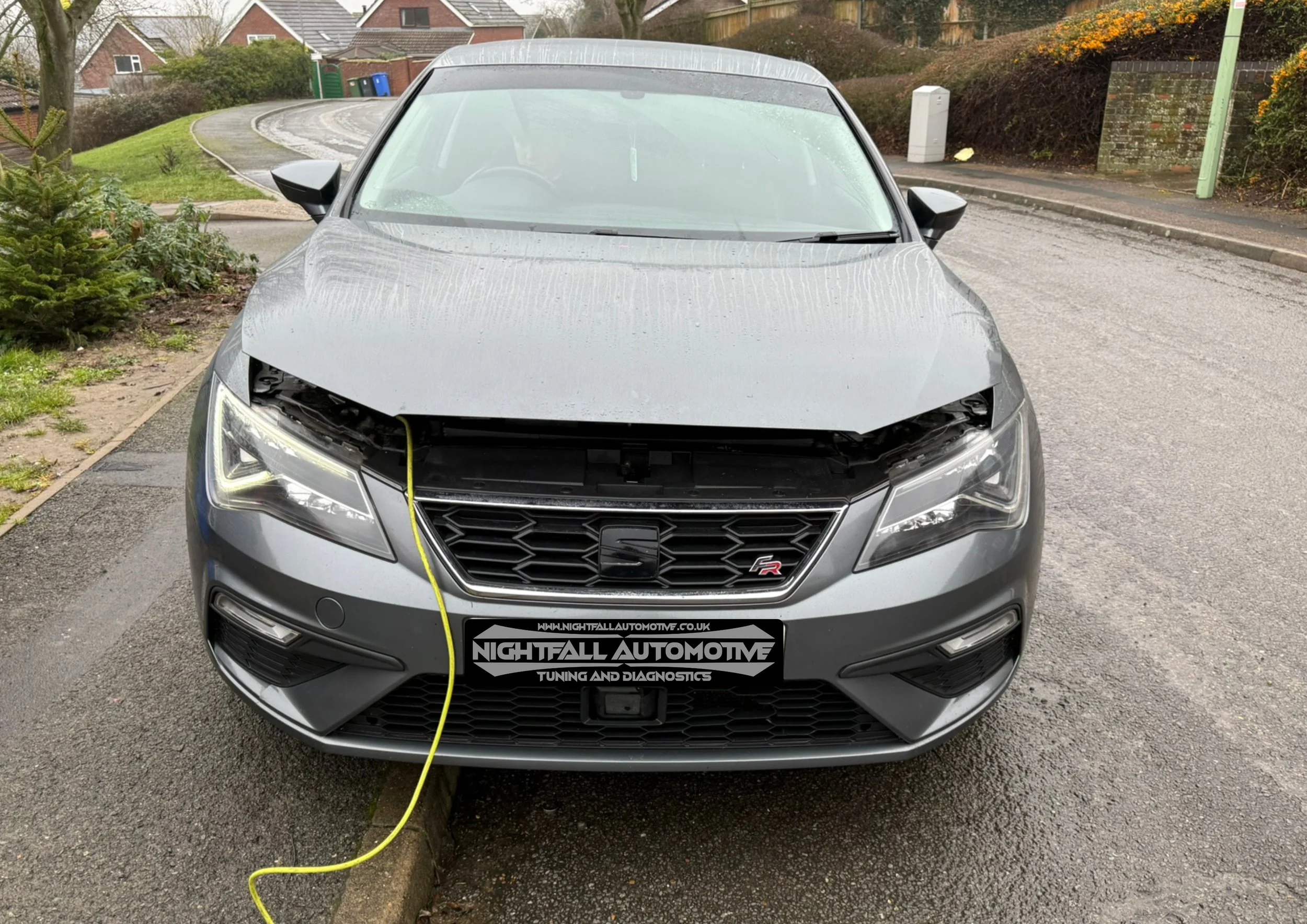 Front view of a silver hatchback car with the front bumper removed, parked on a street with houses and greenery in the background. The car is plugged into a charging station.