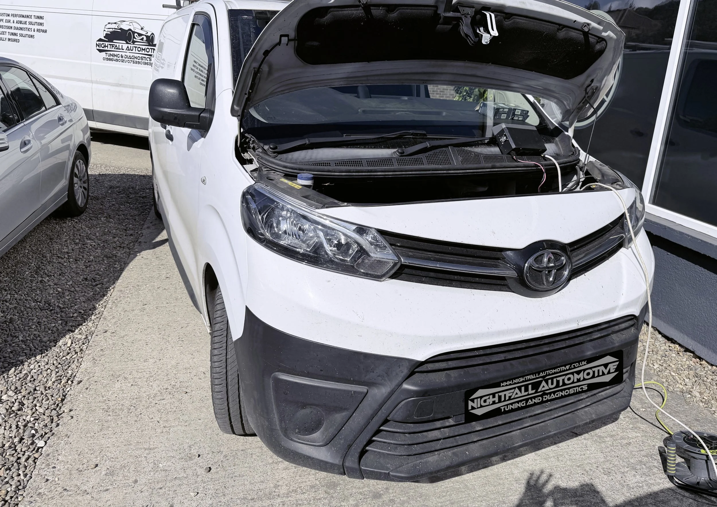 A white compact car with its hood open, parked outside a tuning and diagnostics shop named Nightfall Automotive. The shop's sign is visible on a large white vehicle in the background, Tuned by Nightfall Automotive