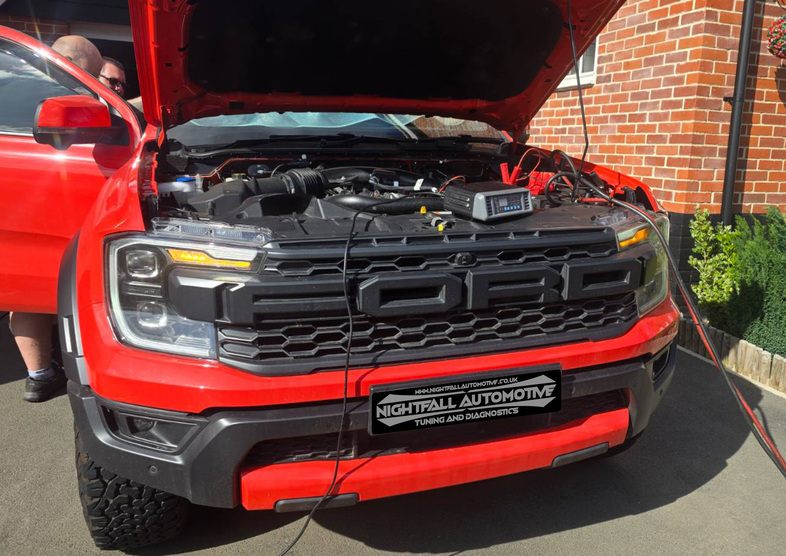 Red Ford truck with open hood, being tested or diagnosed with electronic equipment connected to the engine, parked near a brick house.