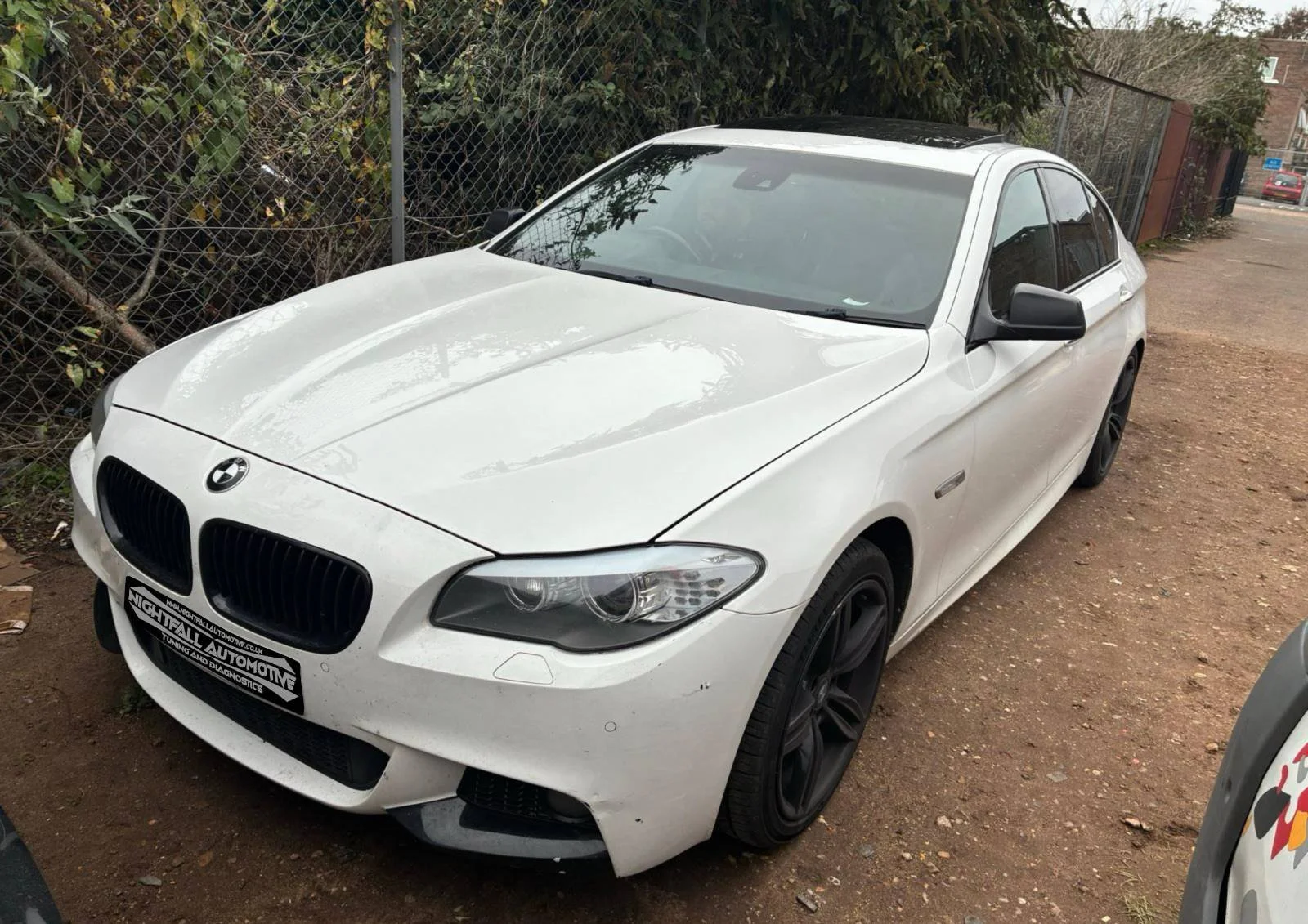 A white BMW sedan parked on a dirt surface next to a chain-link fence with green bushes behind it. The car has black wheels and a black roof, with some dirt visible on the front bumper.