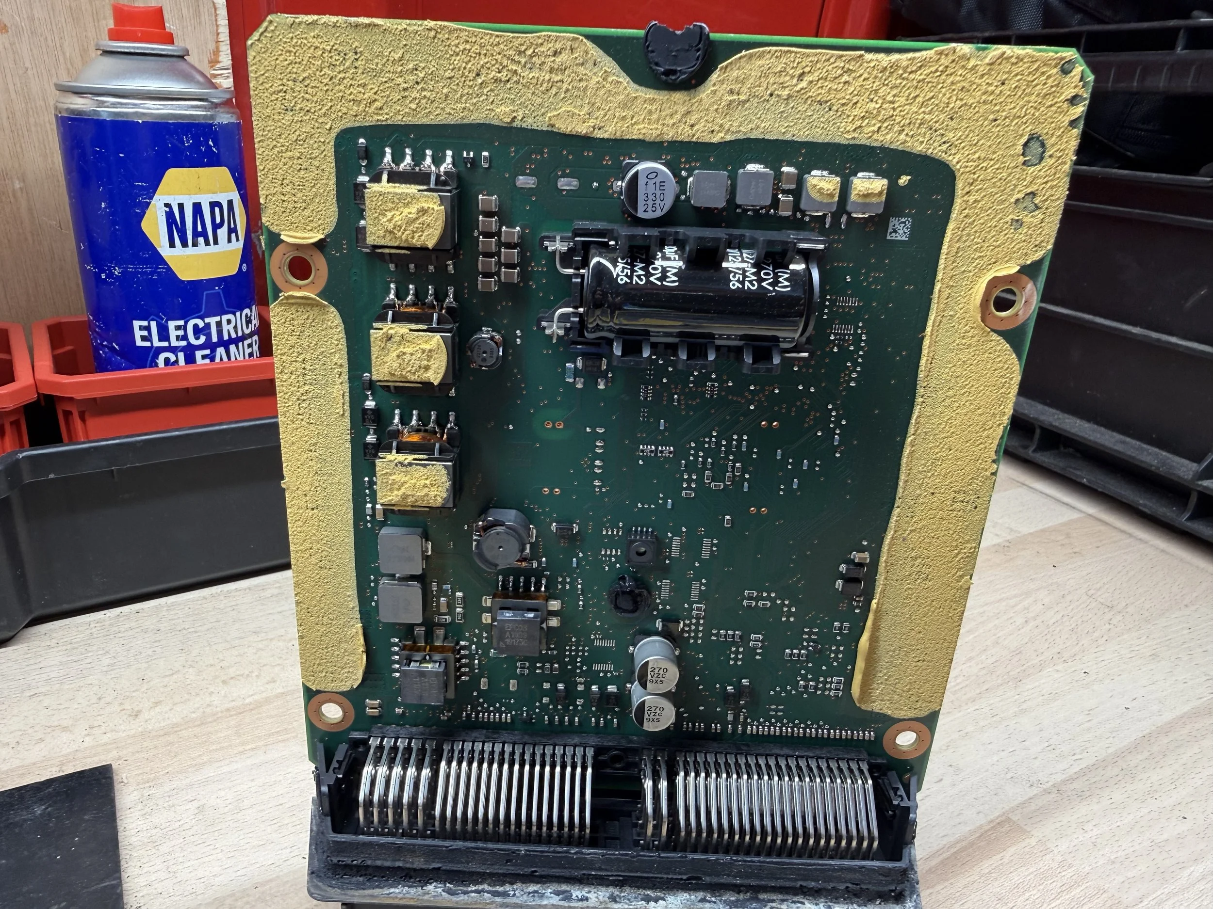 A circuit board with various electronic components, mounted vertically on a work surface, with a can of NAPA electrical cleaner in the background.
