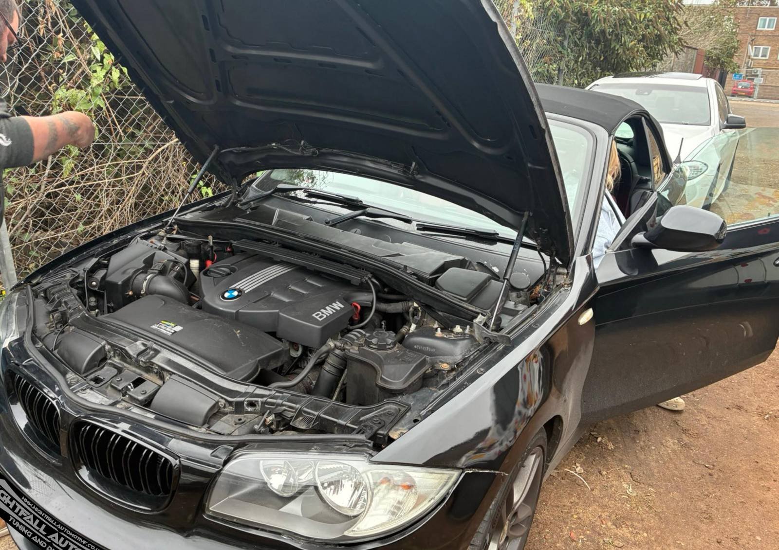 A black BMW convertible with its hood open showing the engine. The car is parked outside on a dirt surface near a chain-link fence, with other vehicles and trees in the background.