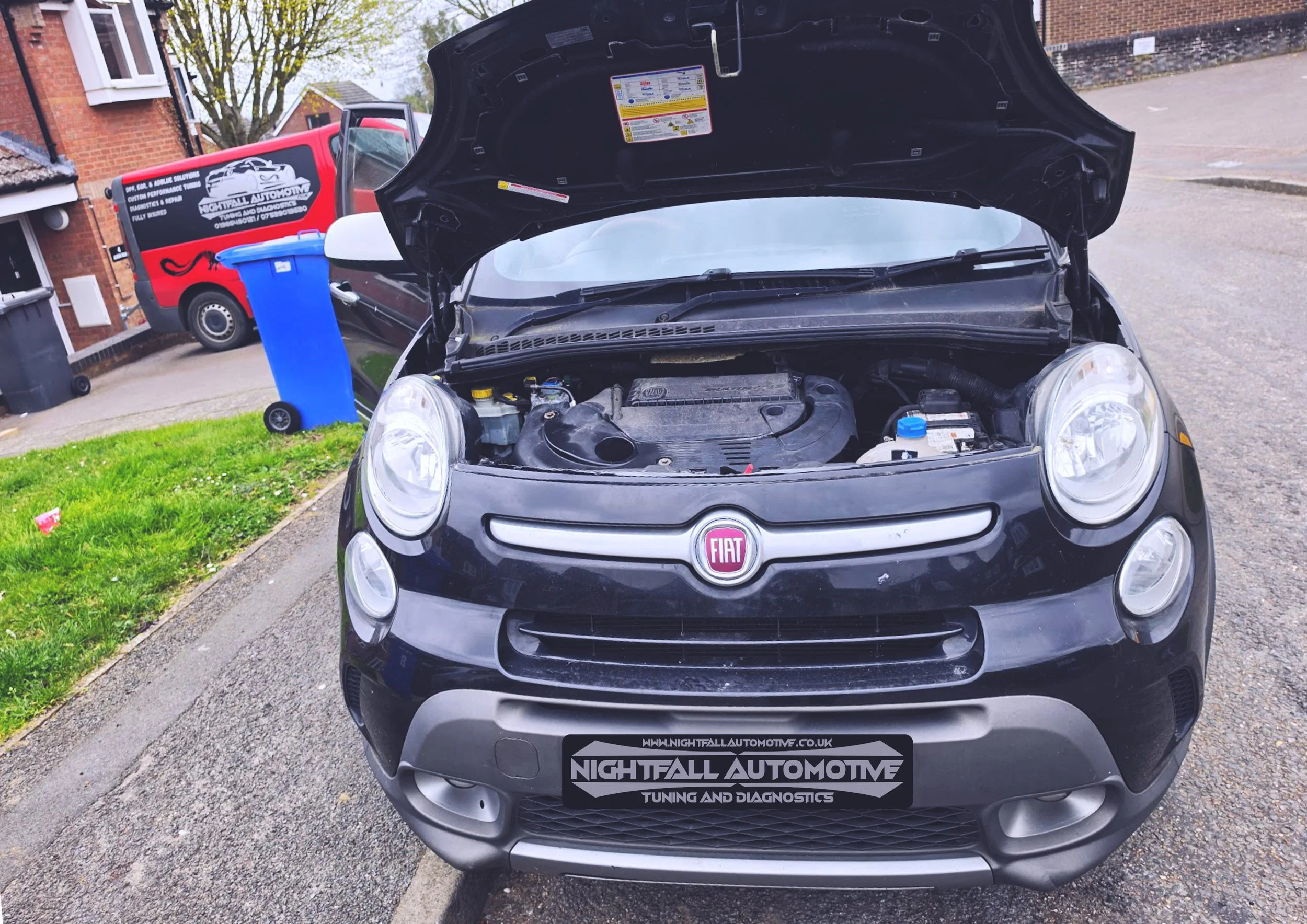 Front view of a small black Fiat car with its hood open, revealing the engine compartment. The car has a Nighfall Automotive tuning and diagnostics sticker on the front bumper. In the background, a red van with advertising for Nighfall Automotive and other vehicles parked nearby in a residential area.