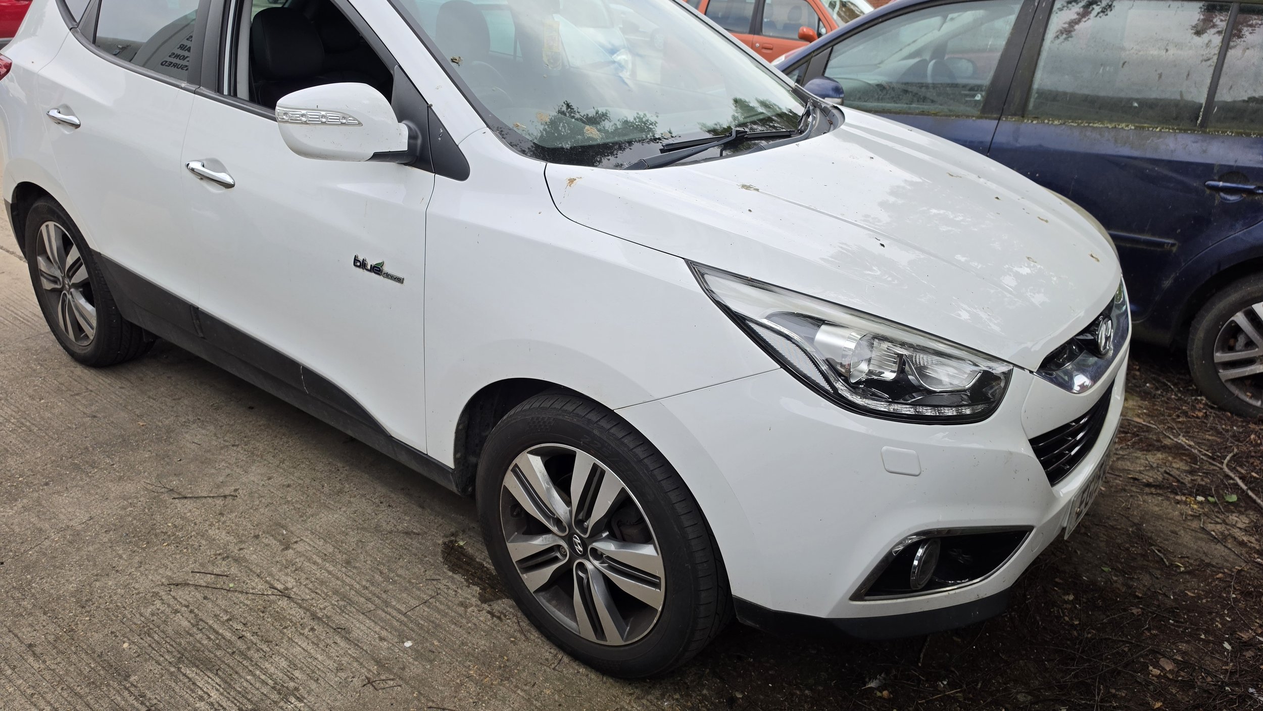 A white electric car parked next to a blue vehicle outside, with a dirt and concrete surface underneath. The white car has a 'blue drive' badge on the side and features modern headlights, side mirrors with LED indicators, and stylish wheels.