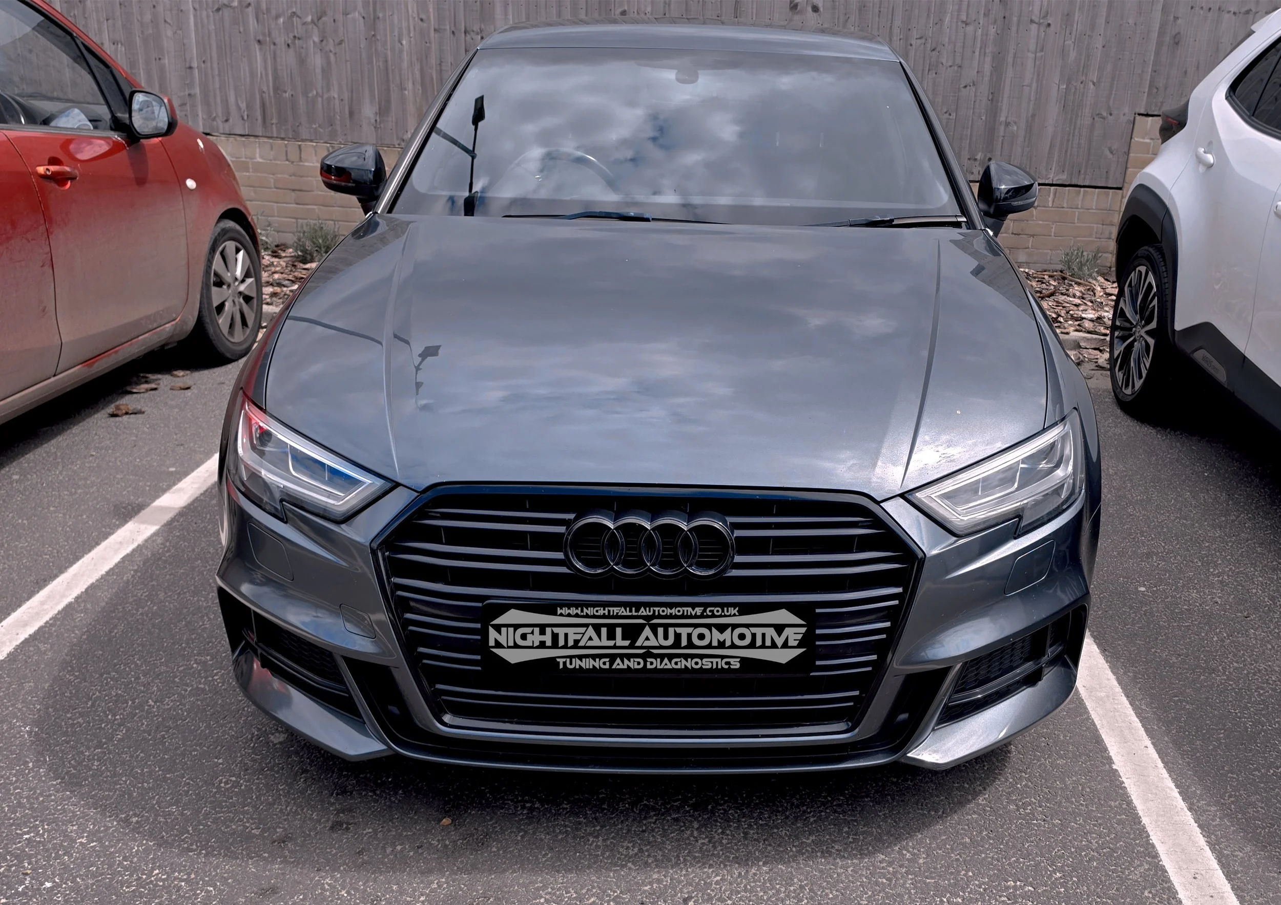 Front view of a gray Audi car parked between a red car on the left and a white car on the right, in a parking lot with a wooden fence background.