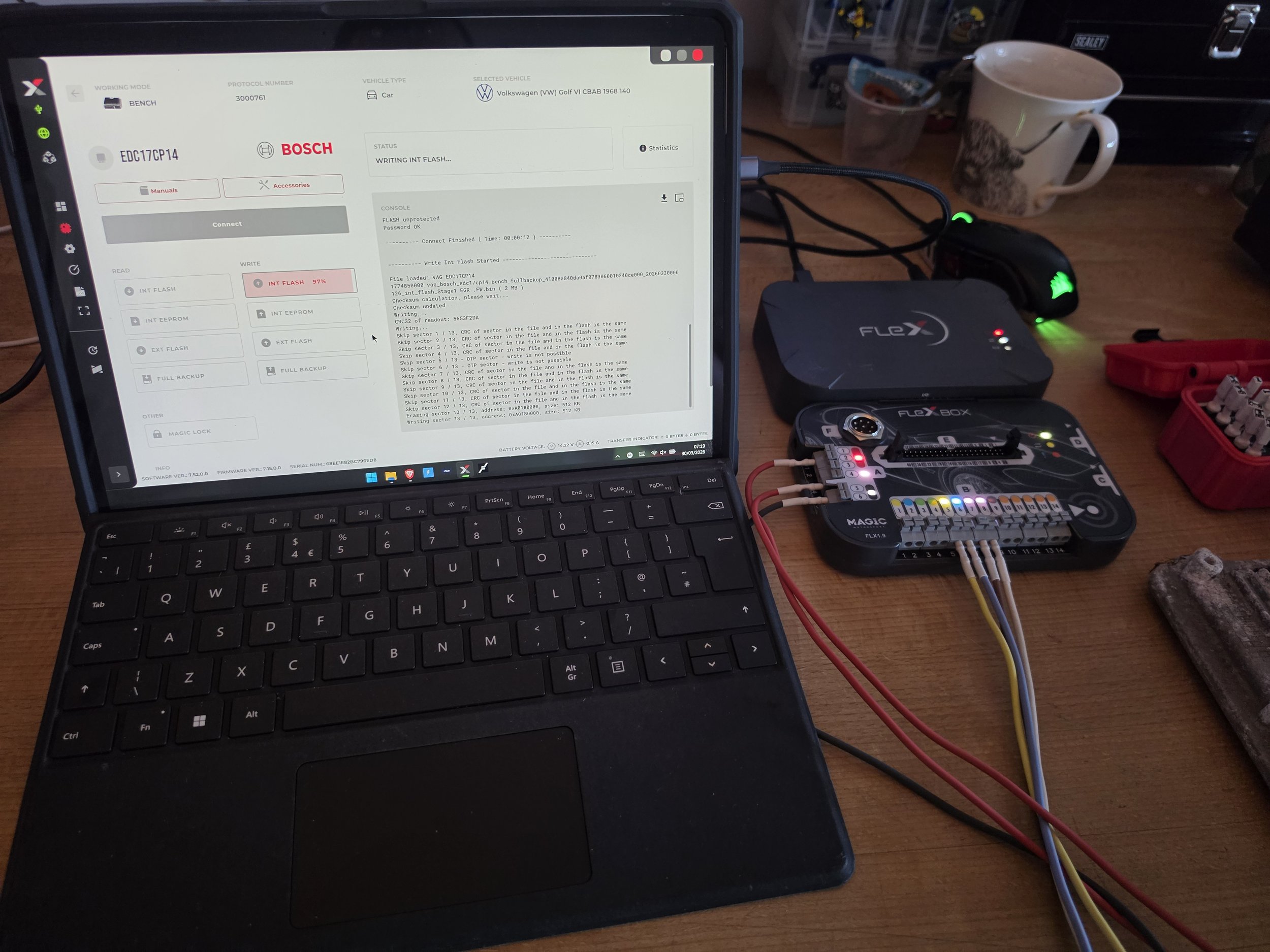 Laptop connected to a hardware programmer and wiring, displaying software interface for vehicle ECU programming on the screen. Items on a wooden desk include a cup, a small red case, Nightfall Automotive DPF Solution