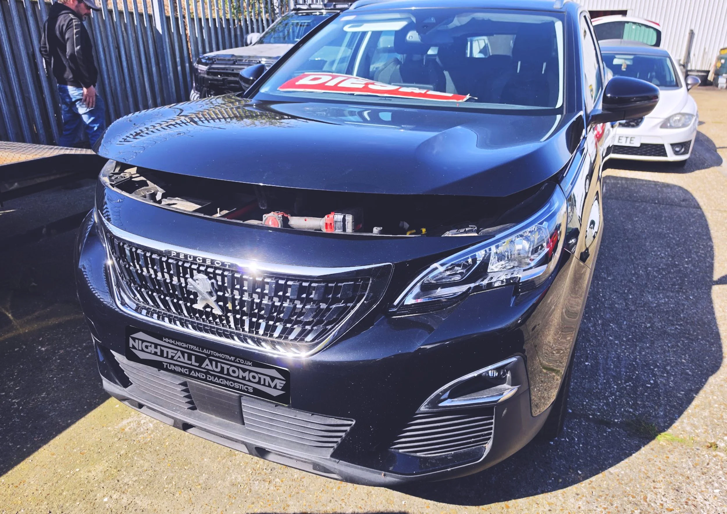 Black Peugeot car with a damaged front, no grille or headlights, parked next to a white car, with a man standing nearby, in an outdoor lot with metal fencing and other vehicles. Nightfall Automotive