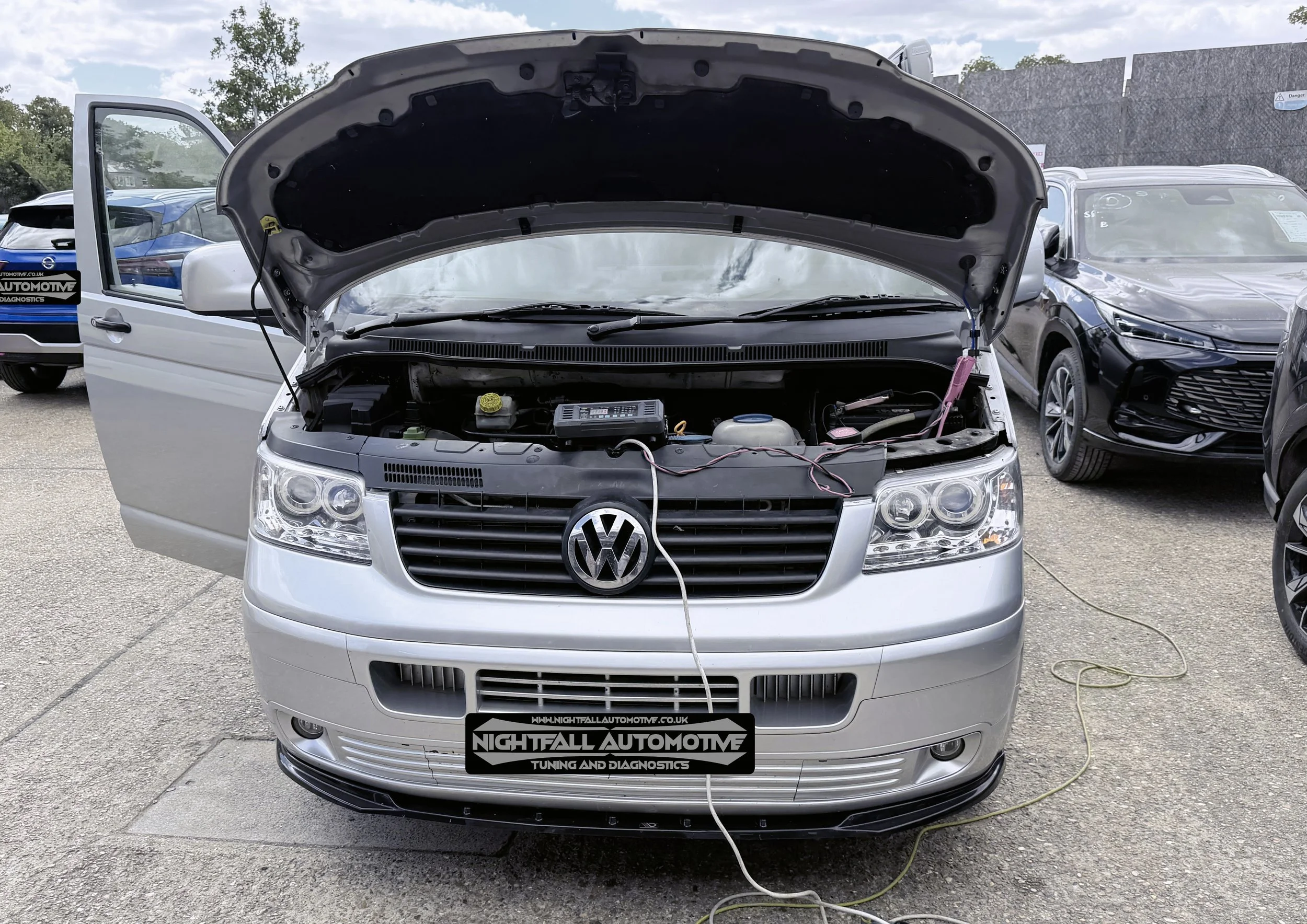 A white Volkswagen vehicle with its hood open, showing the engine compartment, at a car repair or tuning shop with other cars parked nearby.
Tuned by Nightfall Automotive