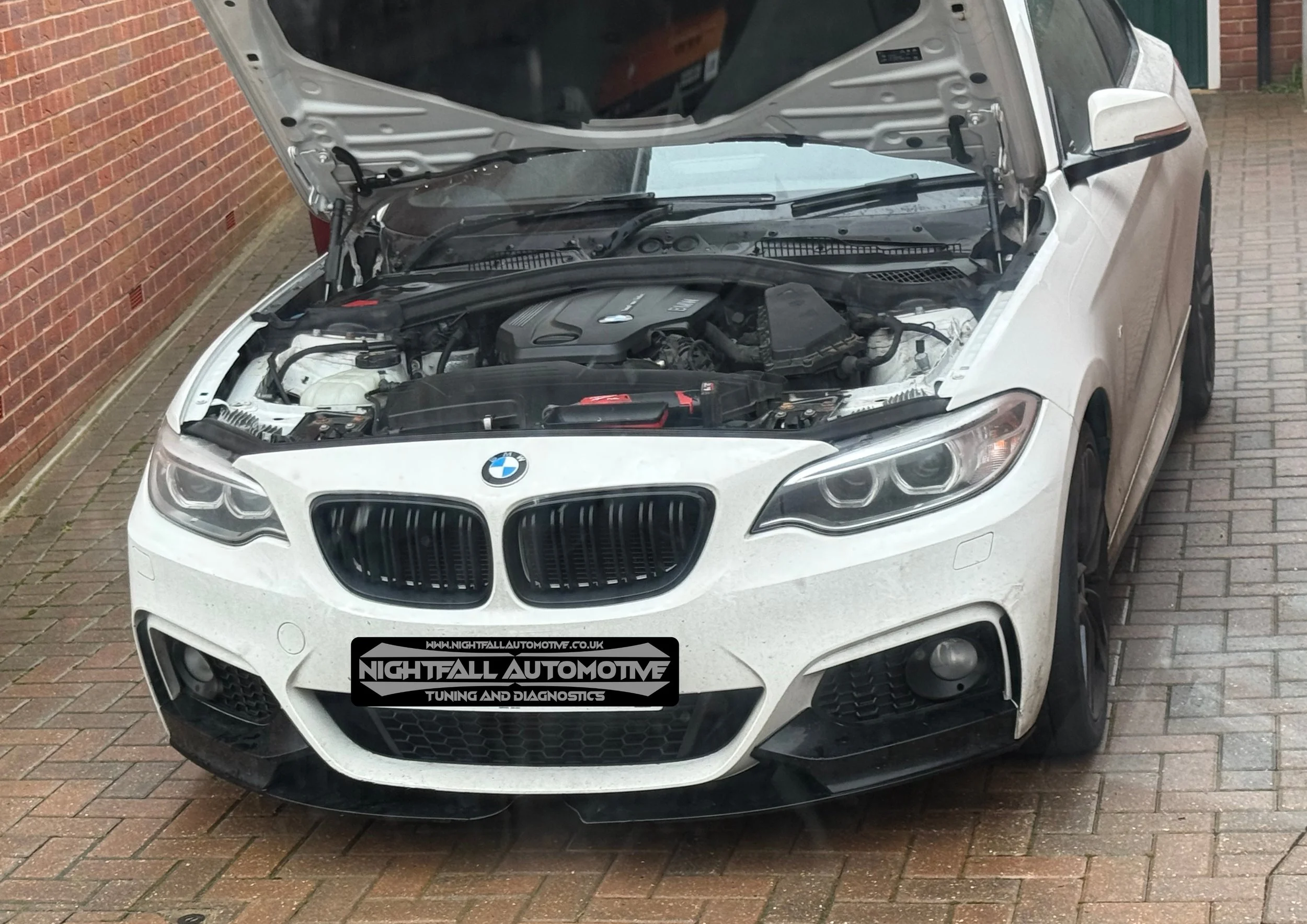 Front view of a white BMW sedan with the hood open, showing the engine bay, parked on a brick driveway next to a brick wall.