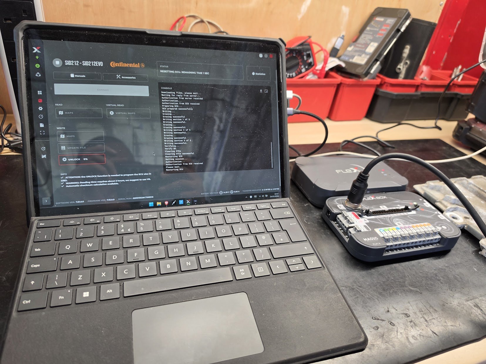 A workbench with a technician's tools, including a tablet displaying software for vehicle ECU programming, a small device connected via a cable, and red plastic bins with additional equipment and wires. work by Nightfall Automotive
