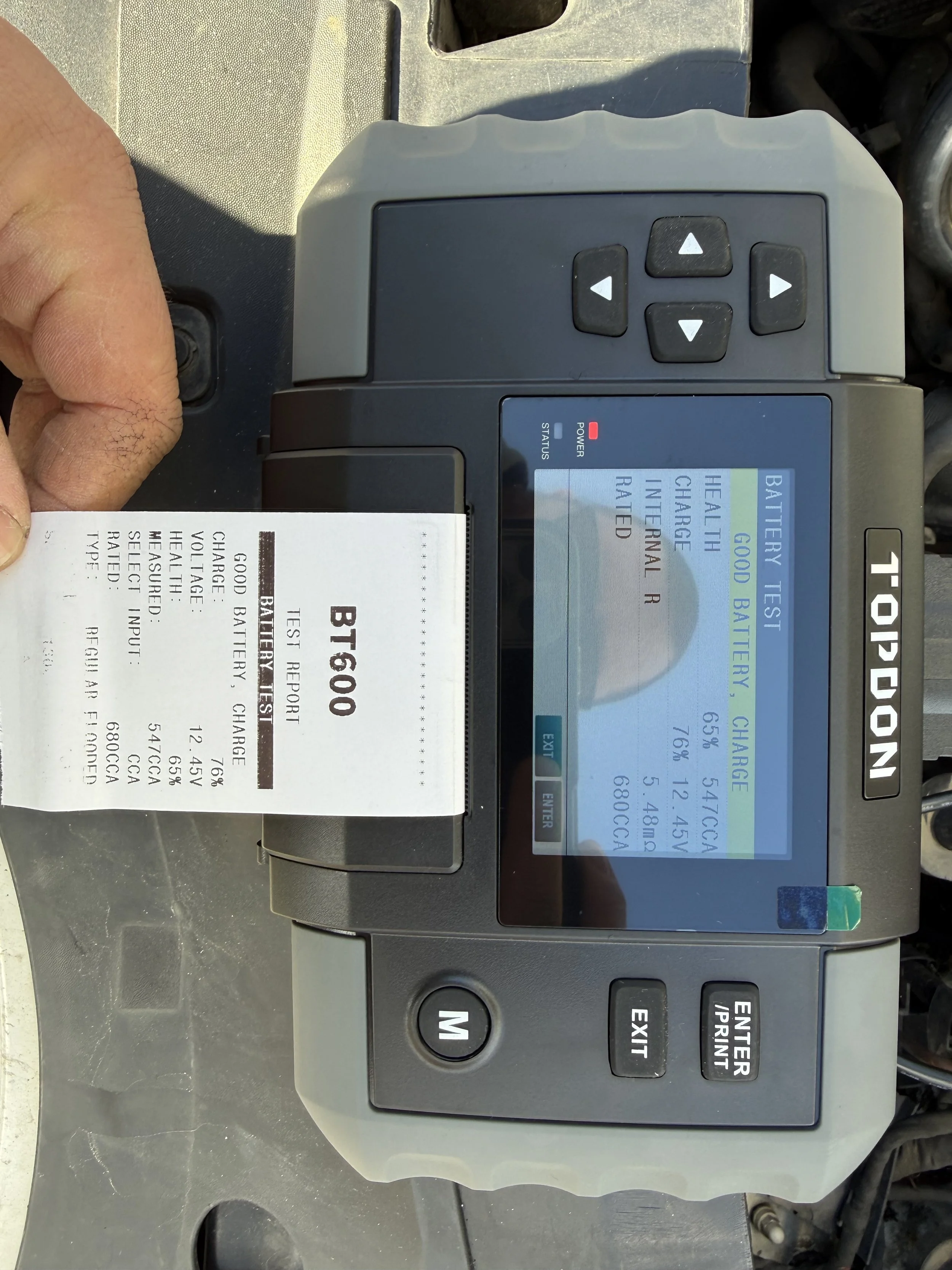 A handheld battery tester connected to a vehicle battery, displaying battery health and charging information on the screen. The device is holding a printout with battery specifications.
