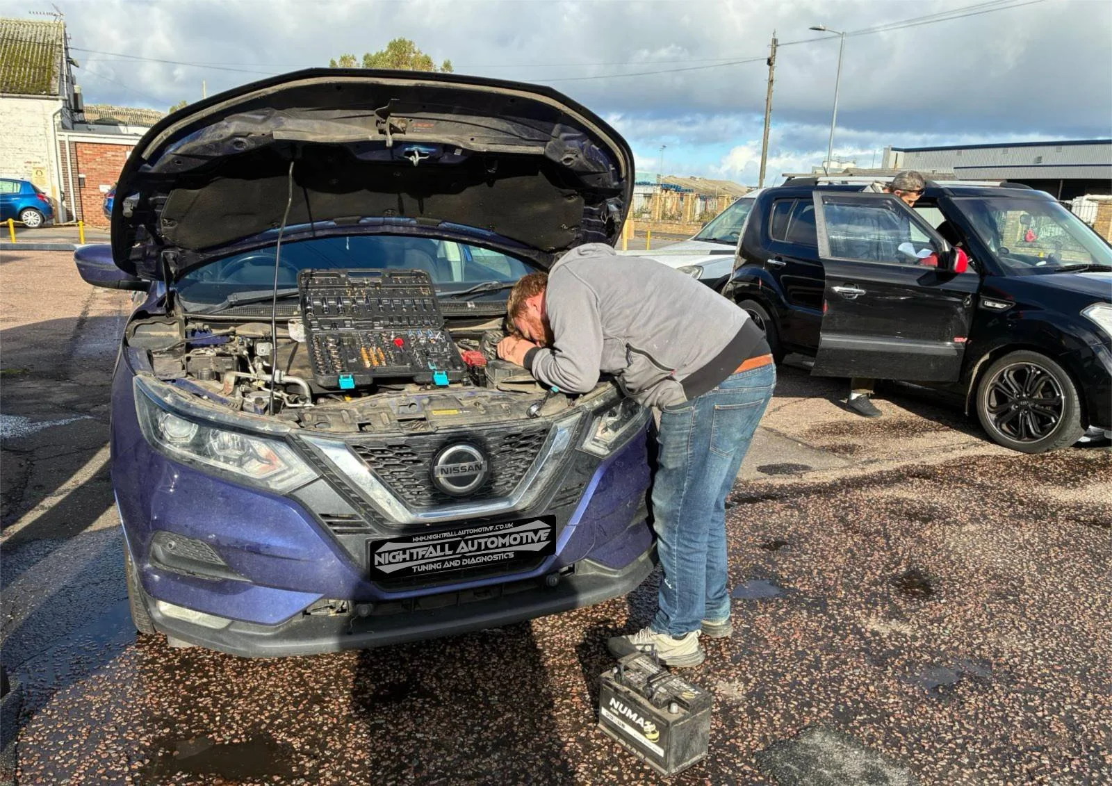 Man inspecting the engine of a purple Nissan car with the hood open in an outdoor parking lot, with vehicles and buildings in the background.