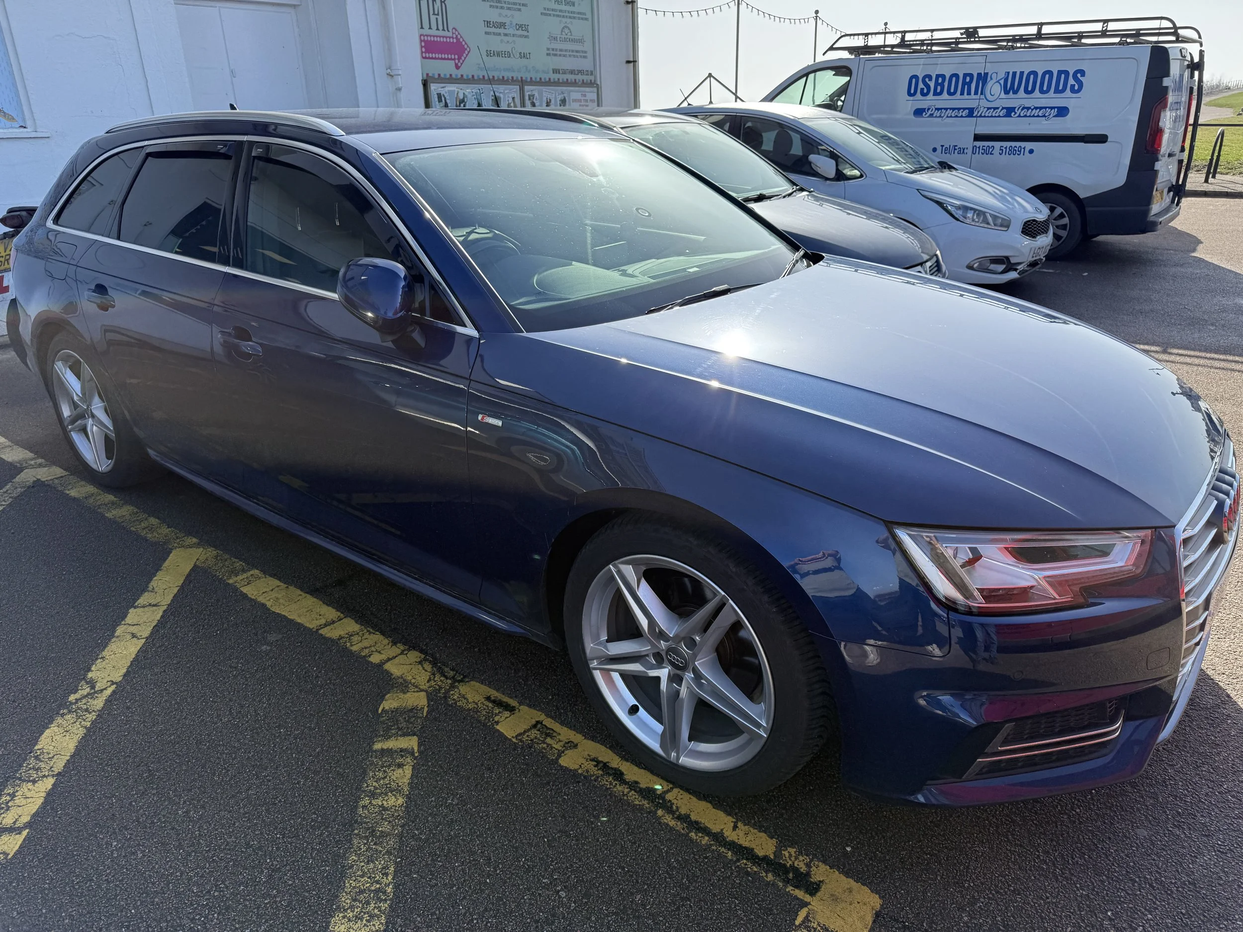 A blue Audi station wagon parked in a lot, with three other cars and a white van in the background. The van has blue text that reads 'Osborn & Woods'.