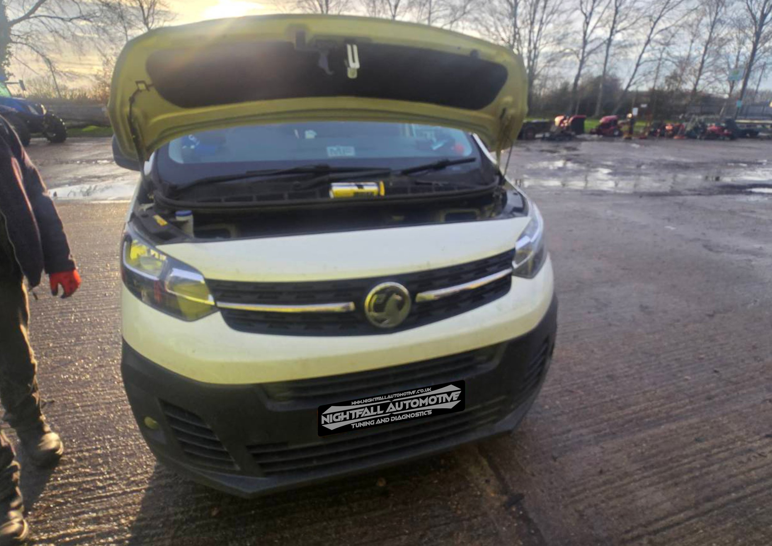 Front view of a white vehicle with its hood open, revealing the engine bay, parked on a muddy lot with other cars in the background.