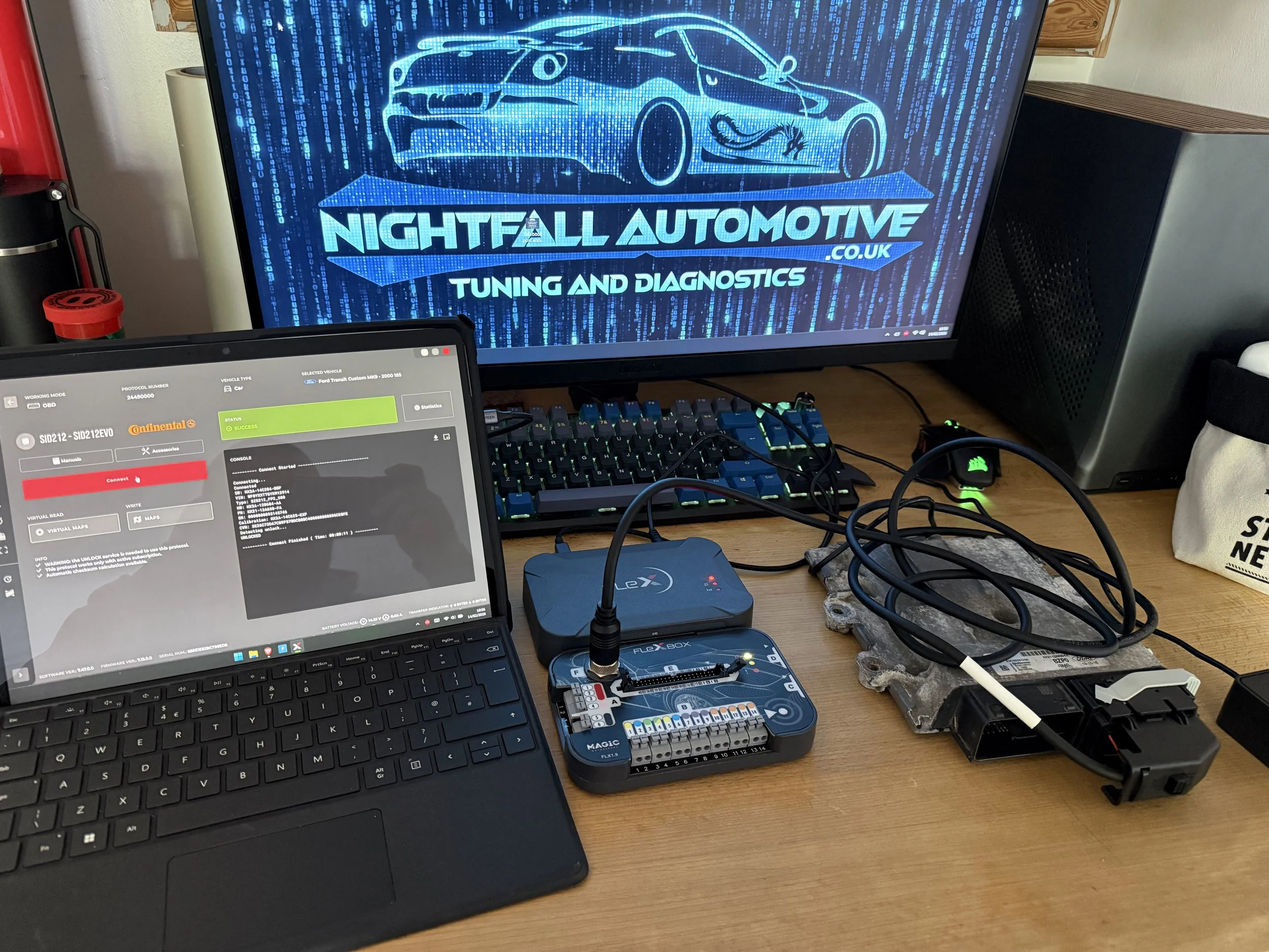 A workstation with a laptop displaying software, a computer monitor with 'Nightfall Automotive Tuning and Diagnostics' logo, and various electronic devices and wiring on a wooden desk.