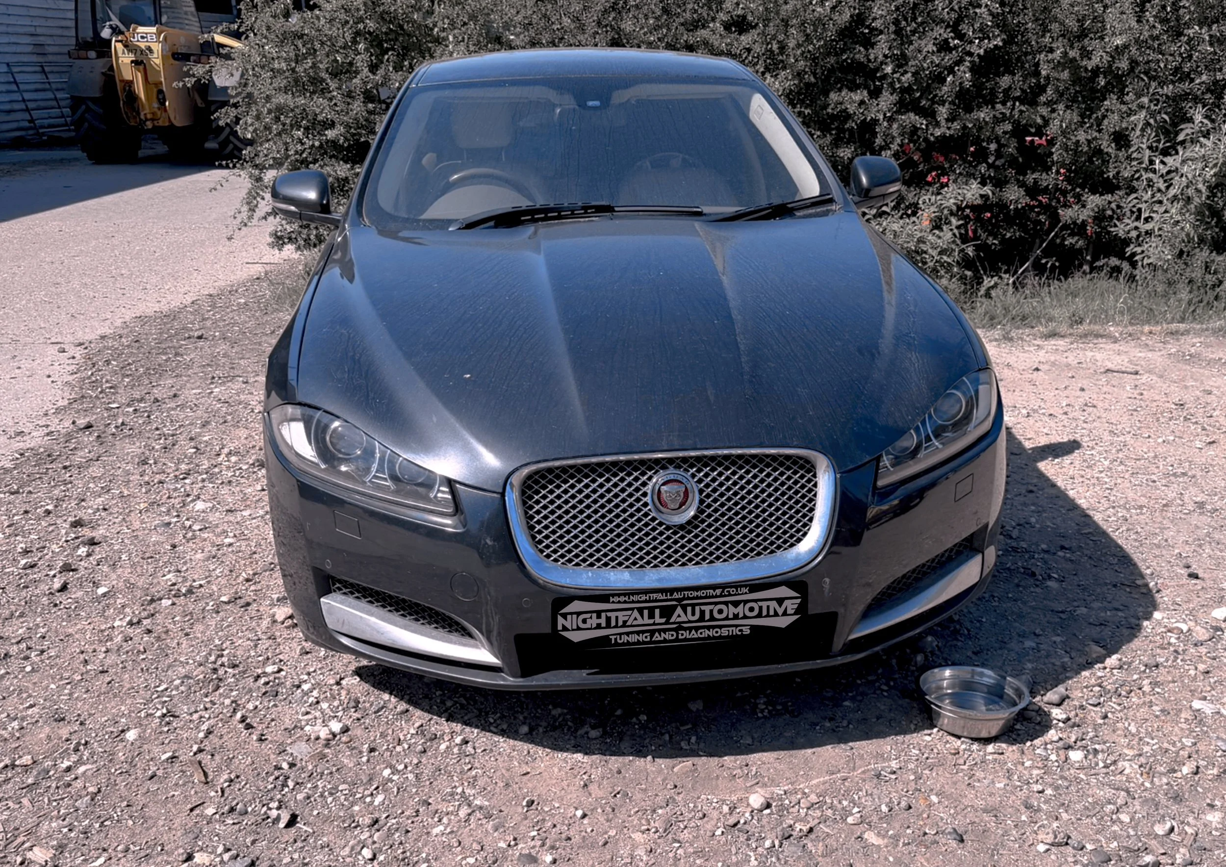 A black Jaguar F-Type parked on a gravel surface with a large bush and construction equipment in the background. There is a metal container on the ground in front of the car. The car's front is visible, showing the grille and headlights.