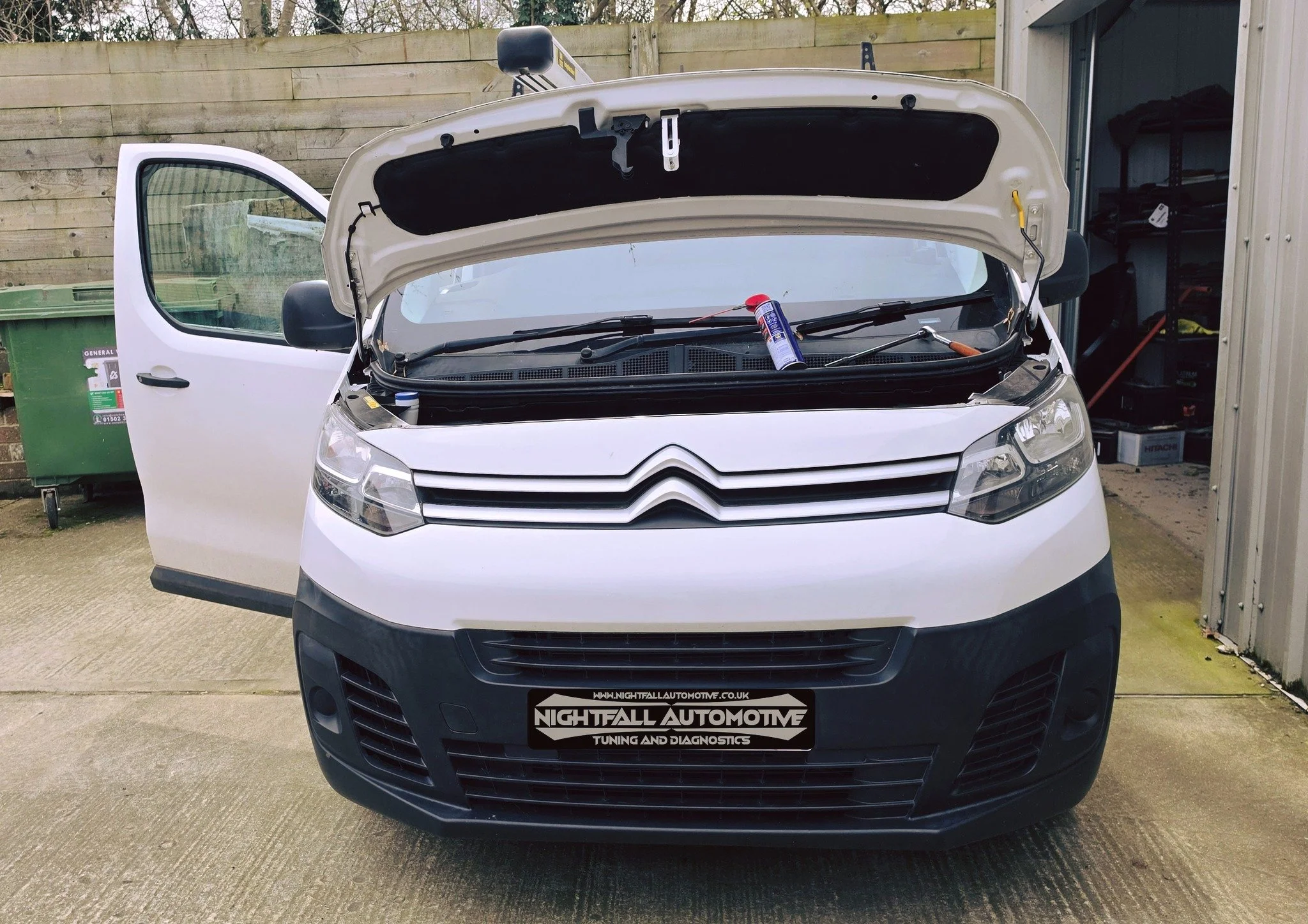 White van with open hood and driver's side door in a driveway, with a bottle of automotive spray inside the engine compartment, a green trash bin nearby, and a shed with tools to the right.