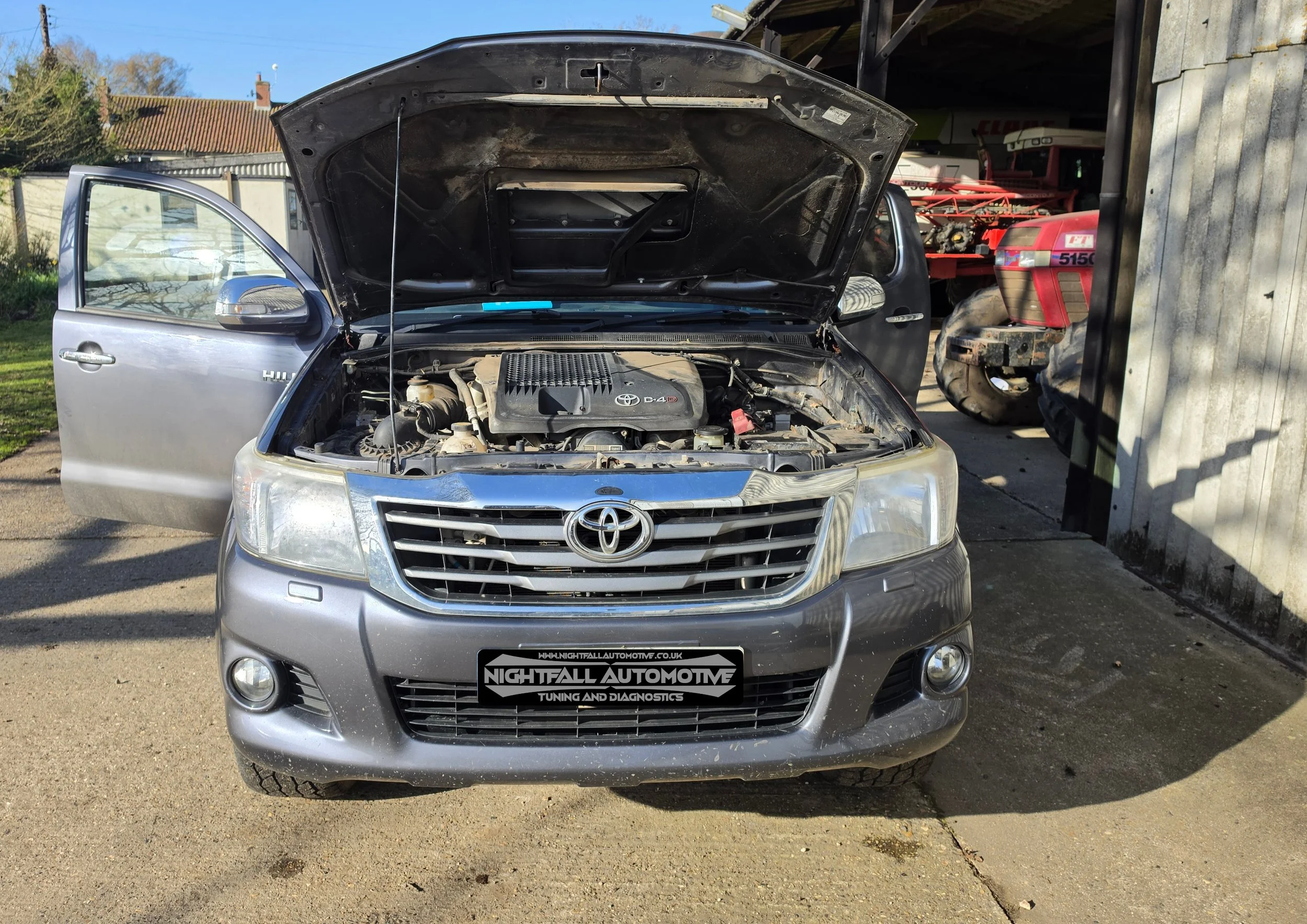 Front view of a silver Toyota SUV with the hood open, showing the engine, parked outside a garage with the driver's door open. Nightfall Automotive stage 1.