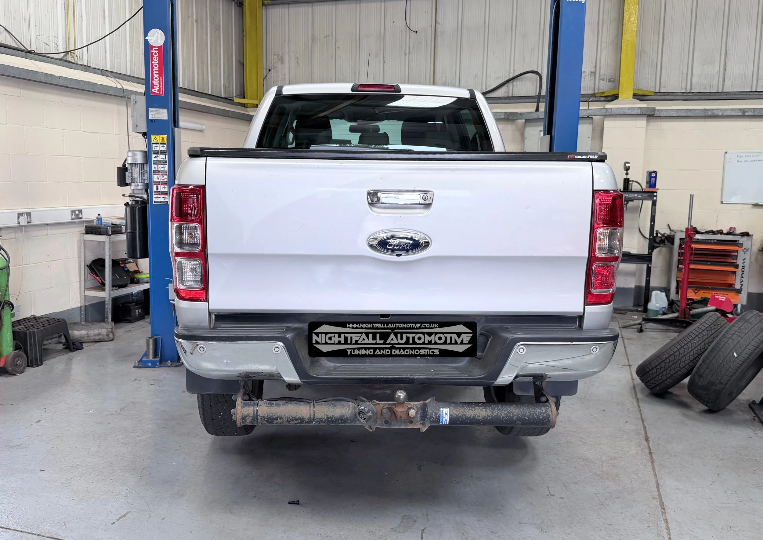 Rear view of a white Ford pickup truck on a vehicle lift inside an auto repair shop. The shop has a concrete floor, with tools and equipment visible around. Tuned by Nightfall Automotive