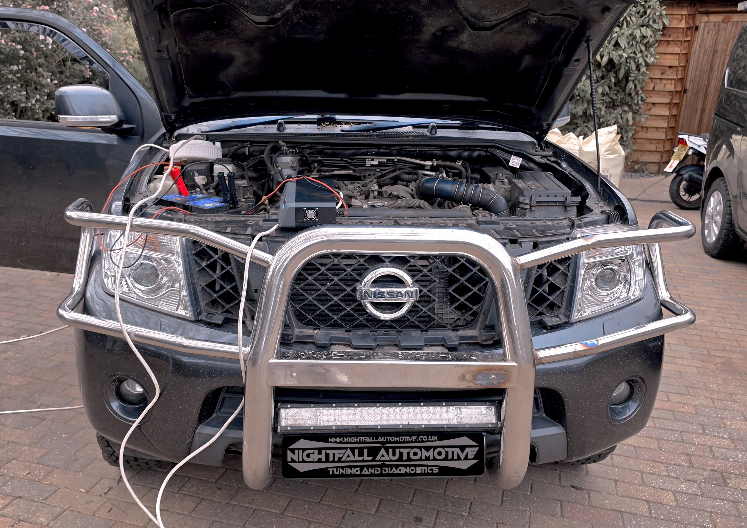 A black Nissan vehicle with its hood open, showing the engine compartment. There is a metal grill guard on the front, and the vehicle is parked on a paved driveway. A sign on the front bumper reads 'Nightfall Automotive Tuning and Diagnostics'.