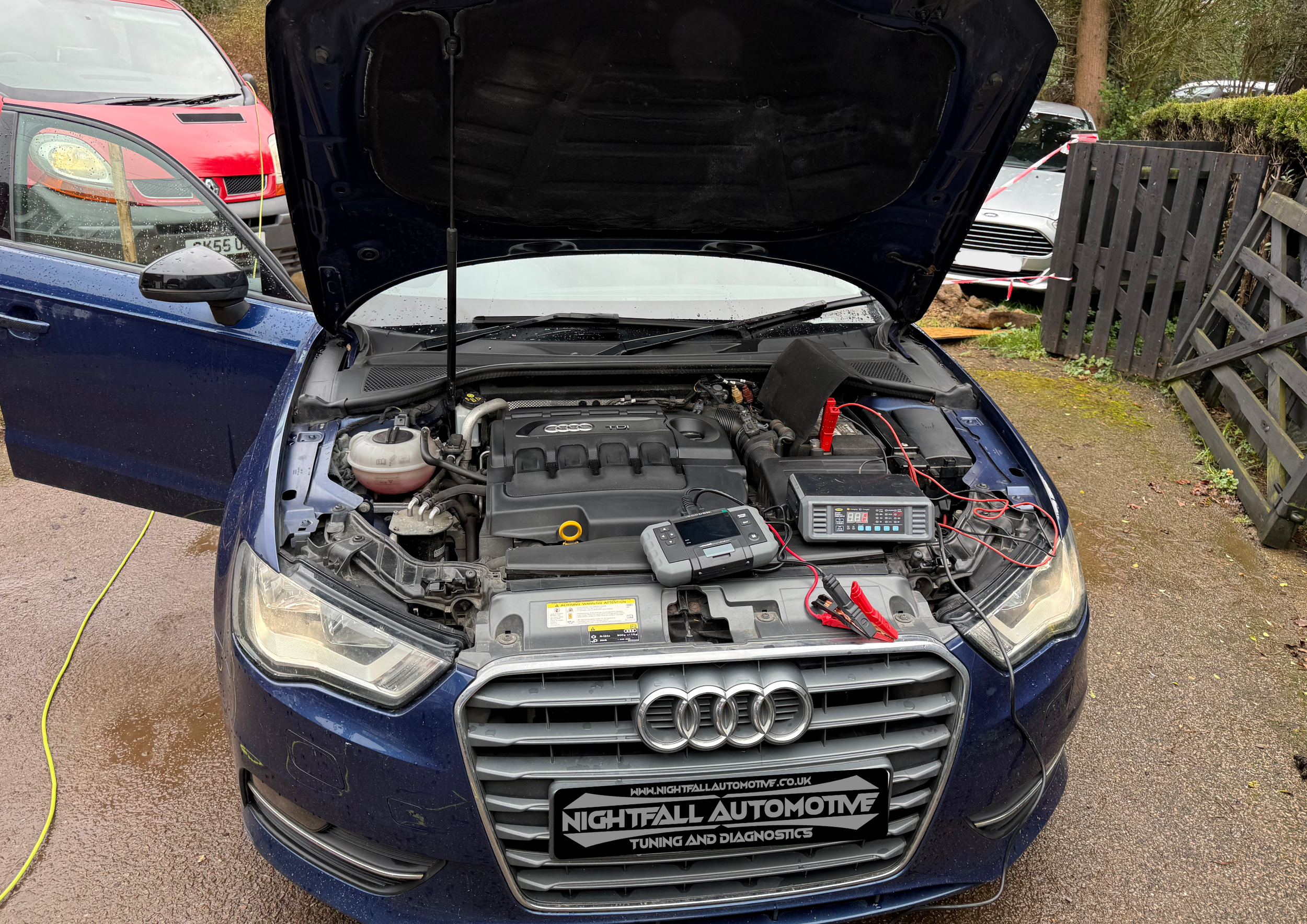 An Audi car undergoing tuning and diagnostics with its hood open, connected to testing equipment. The car is parked outdoors on a wet surface with other cars and a wooden fence in the background.