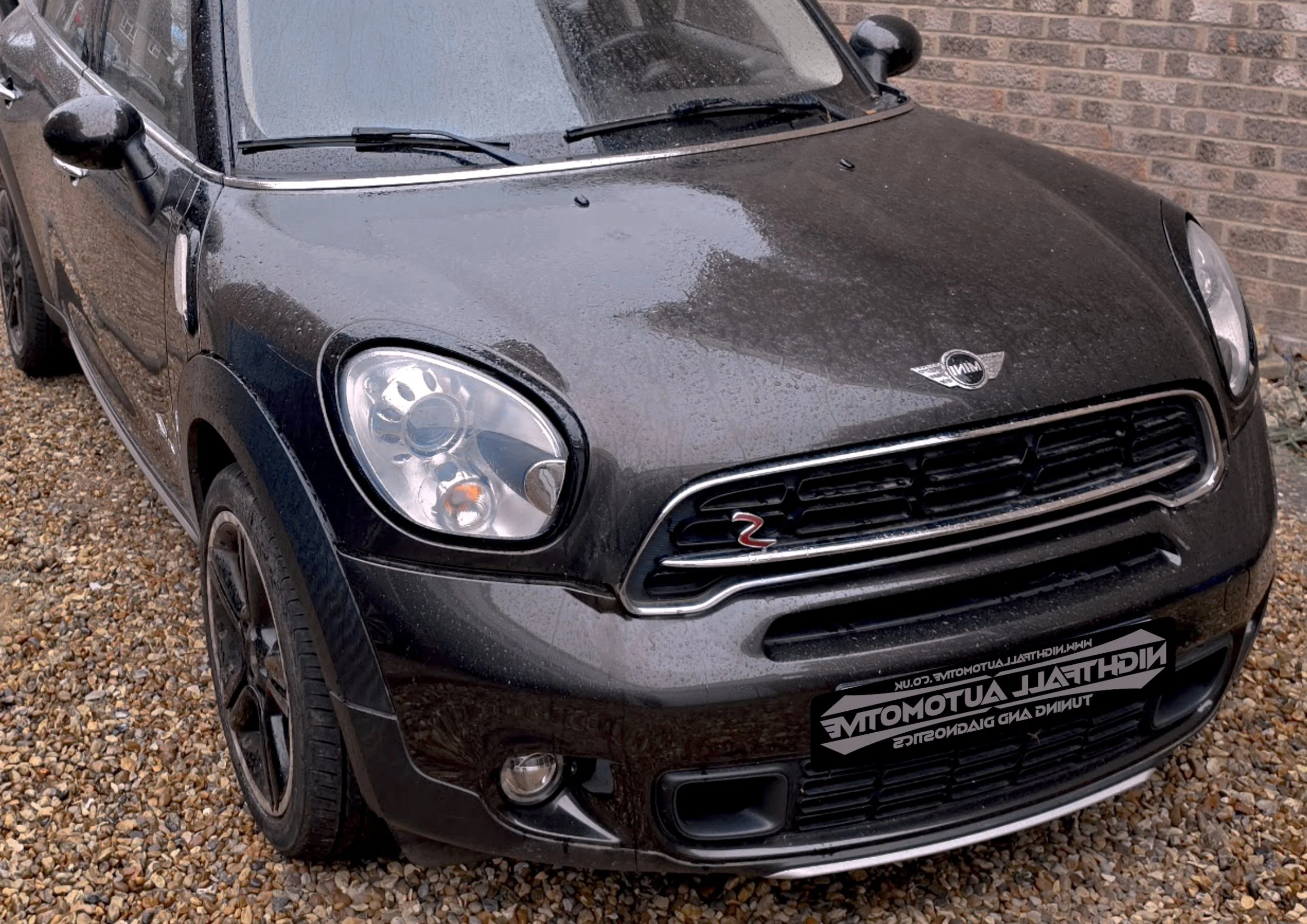 A black Mini Cooper car with rain droplets on its surface, parked on a gravel surface near a brick wall. tuned by Nightfall Automotive