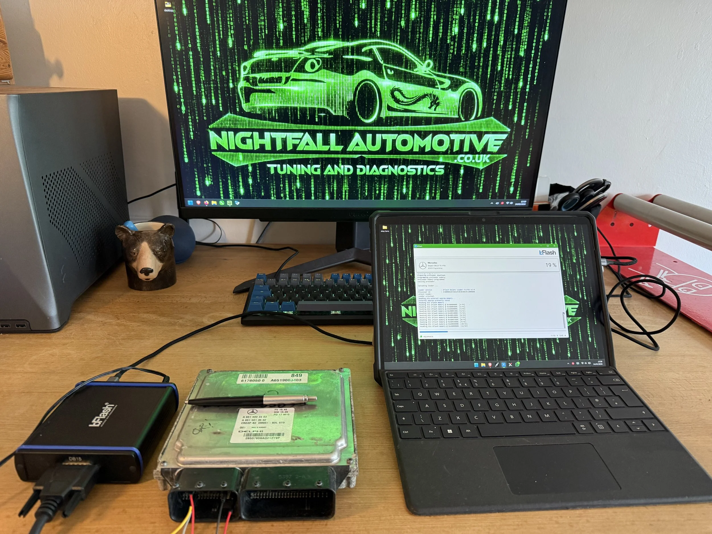 Workstation setup with a monitor displaying the logo for Nightfall Automotive, a tablet with a green digital background, a desktop computer, a hard drive, a pen, and a small bear-shaped figurine on the wooden desk.