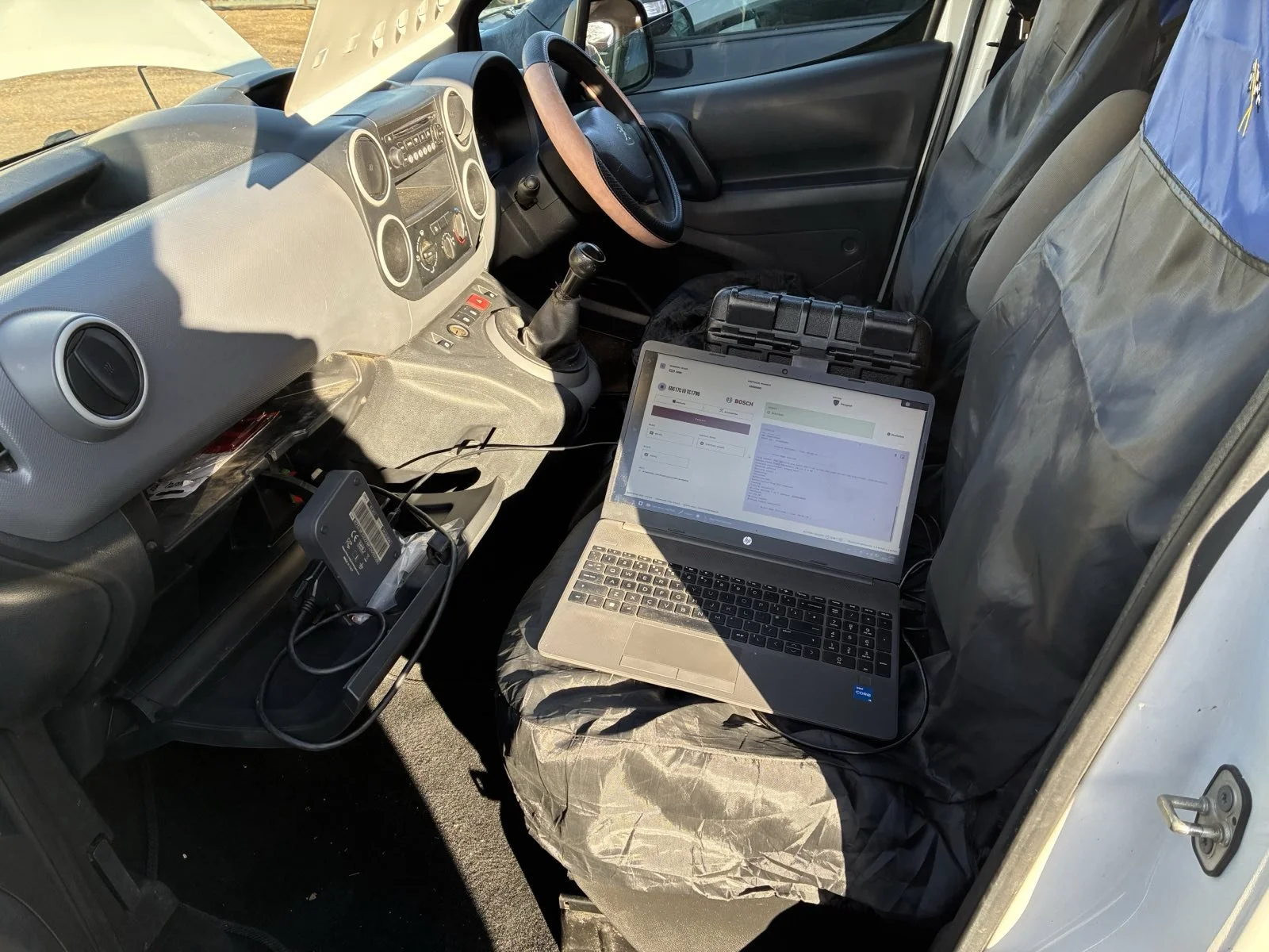 Inside a vehicle, the driver’s seat and dashboard are visible, with a laptop, diagnostic device, and cables connected to the vehicle. The laptop screen displays diagnostic software.