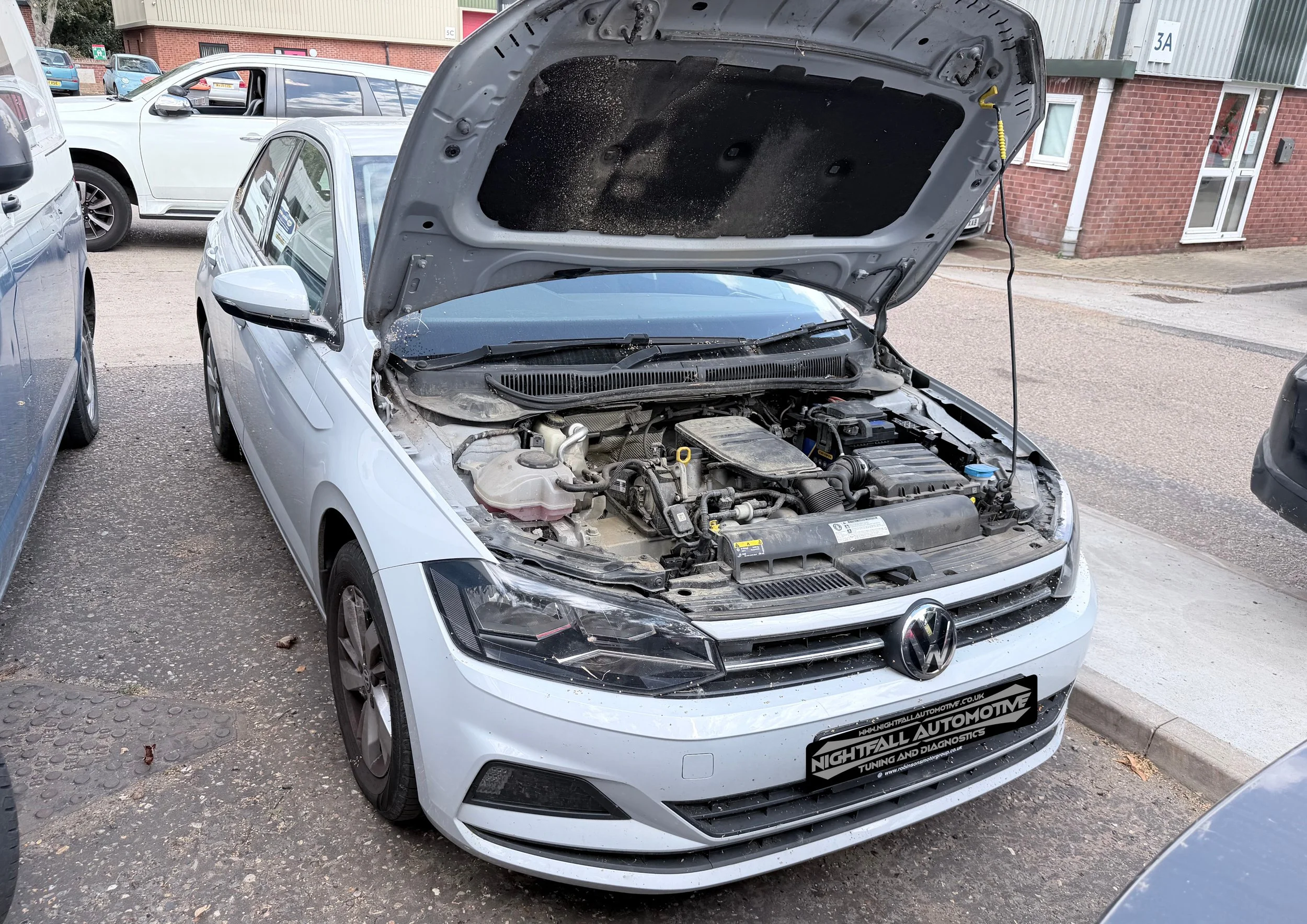 A silver Volkswagen car with its hood open, parked on a street with other vehicles and buildings in the background. Tuned by Nightfall Automotive
