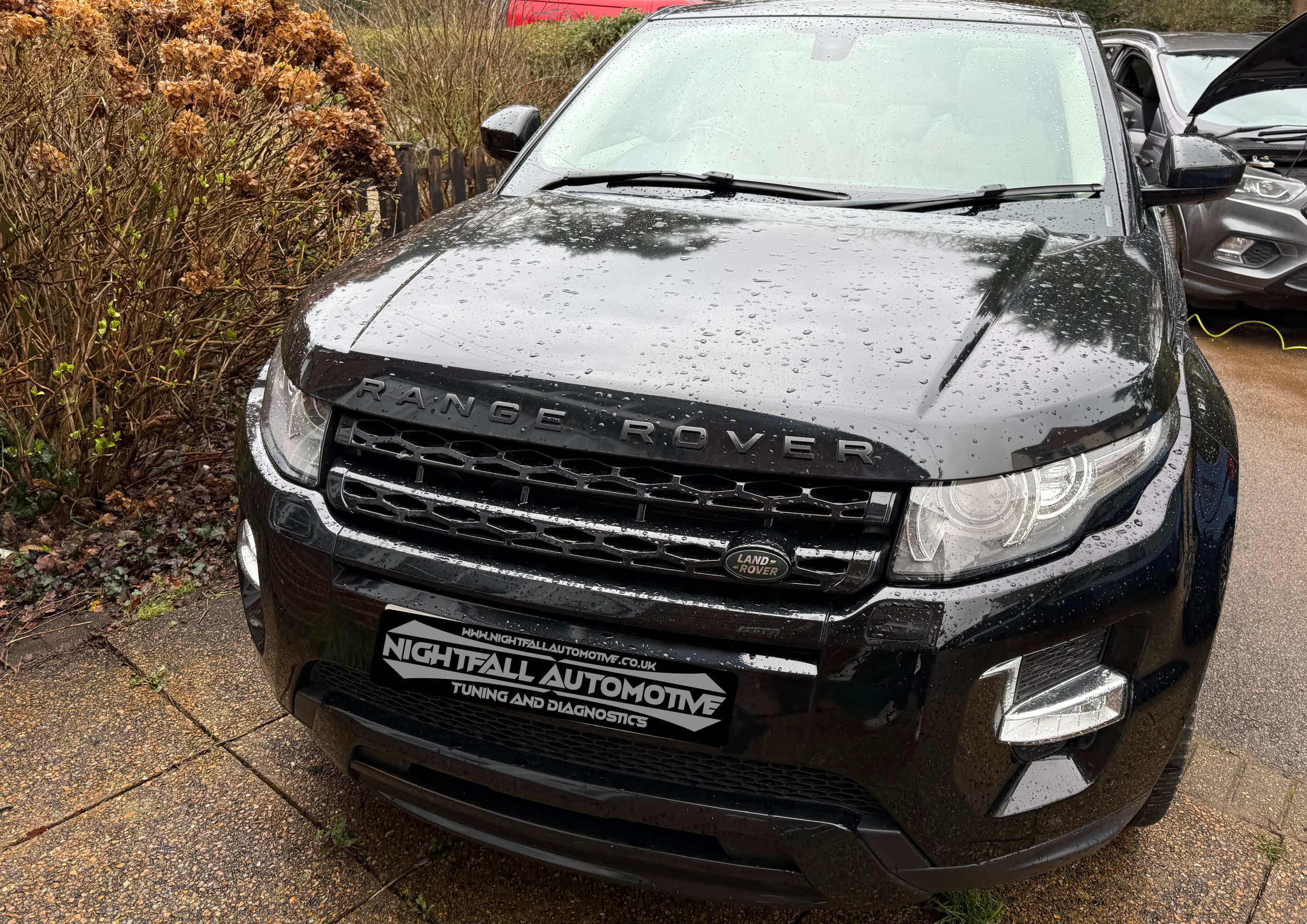 Black Range Rover with raindrops on the hood, parked on a brick pavement next to bushes. The front grille has a Land Rover badge and a license plate advertising Nightfall Automotive, which offers tuning and diagnostics.