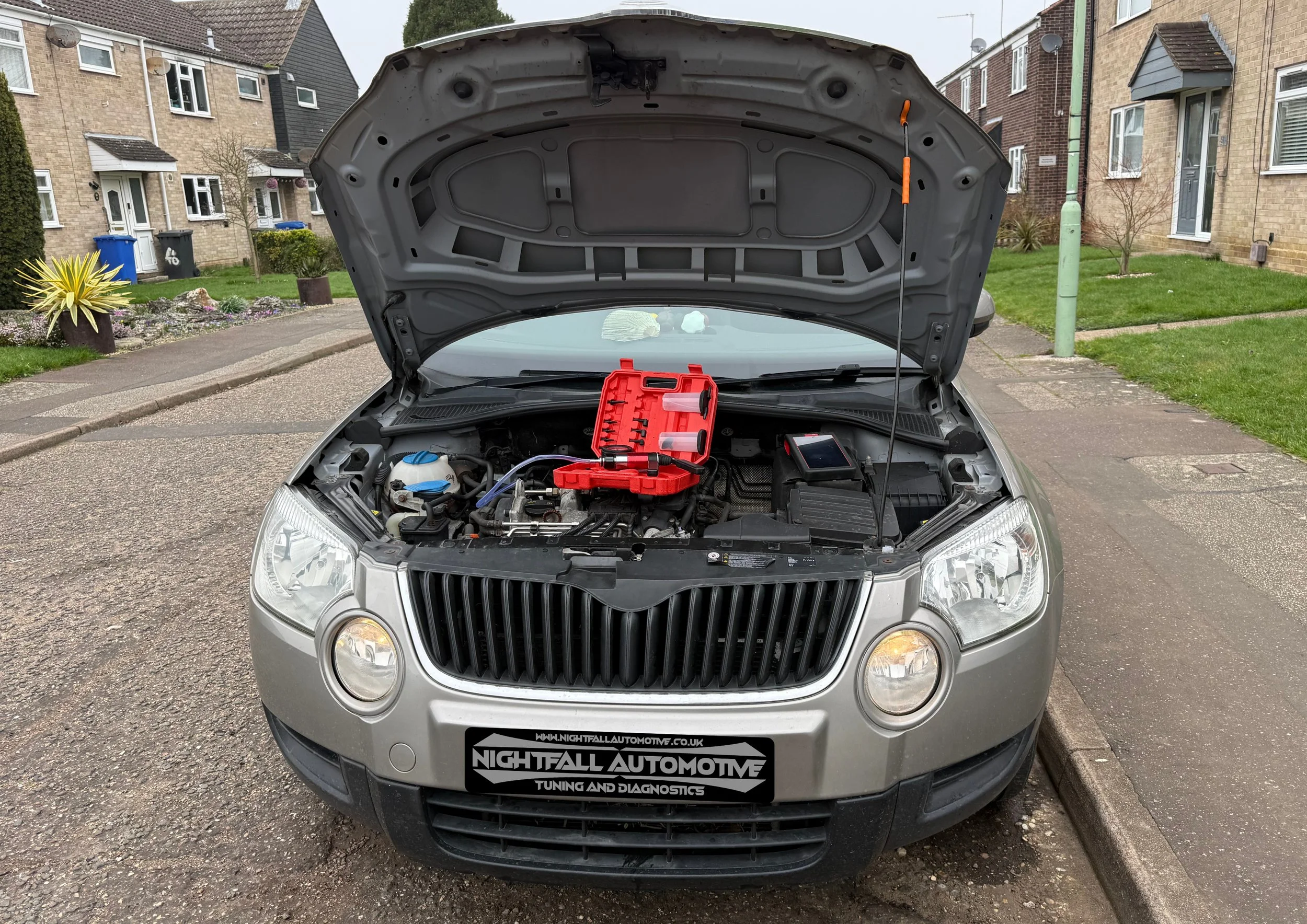 A silver car with its hood open, showing the engine bay with a red toolbox resting on top of the engine. The car is parked on a residential street with houses and greenery in the background.