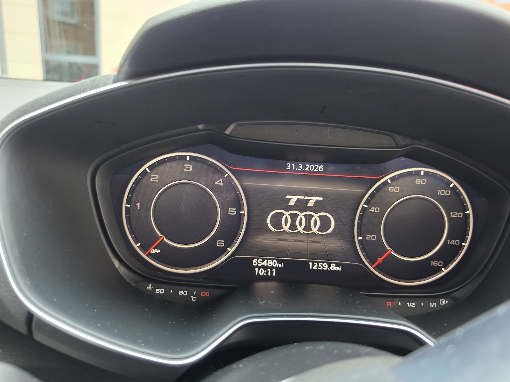 The digital car dashboard displays the Audi TT logo with Audi symbol, showing the date 31.3.2026, the odometer reading 65,480 miles, the trip distance 1,259.8 miles, and the time 10:11. The left dial shows engine RPM near 0, and the right dial shows 