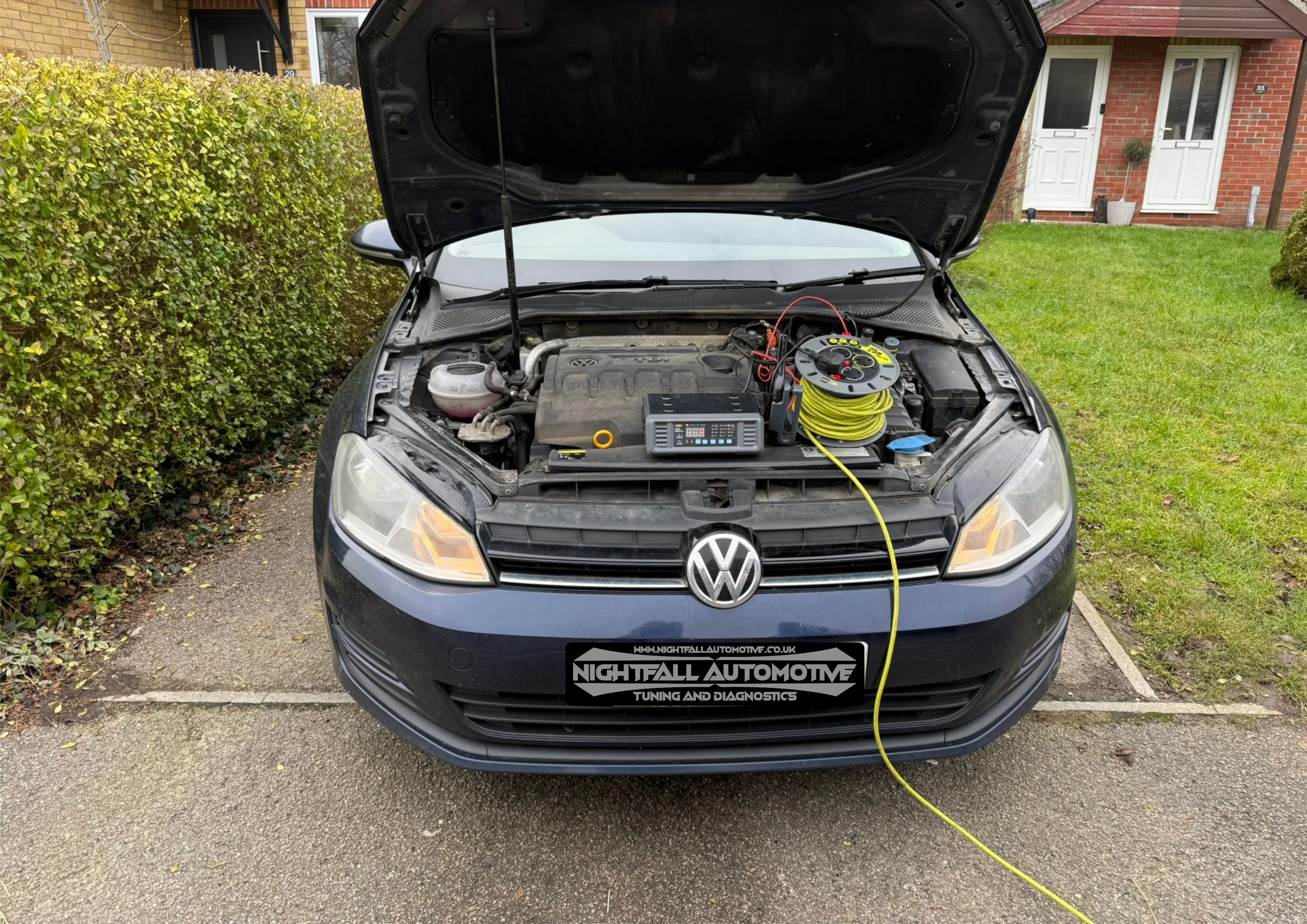 Front view of a black Volkswagen car with the hood open, showing the engine and diagnostic tools placed on top. The car is parked on a driveway next to a grassy area and a hedge, with residential buildings in the background. A yellow cable is connect