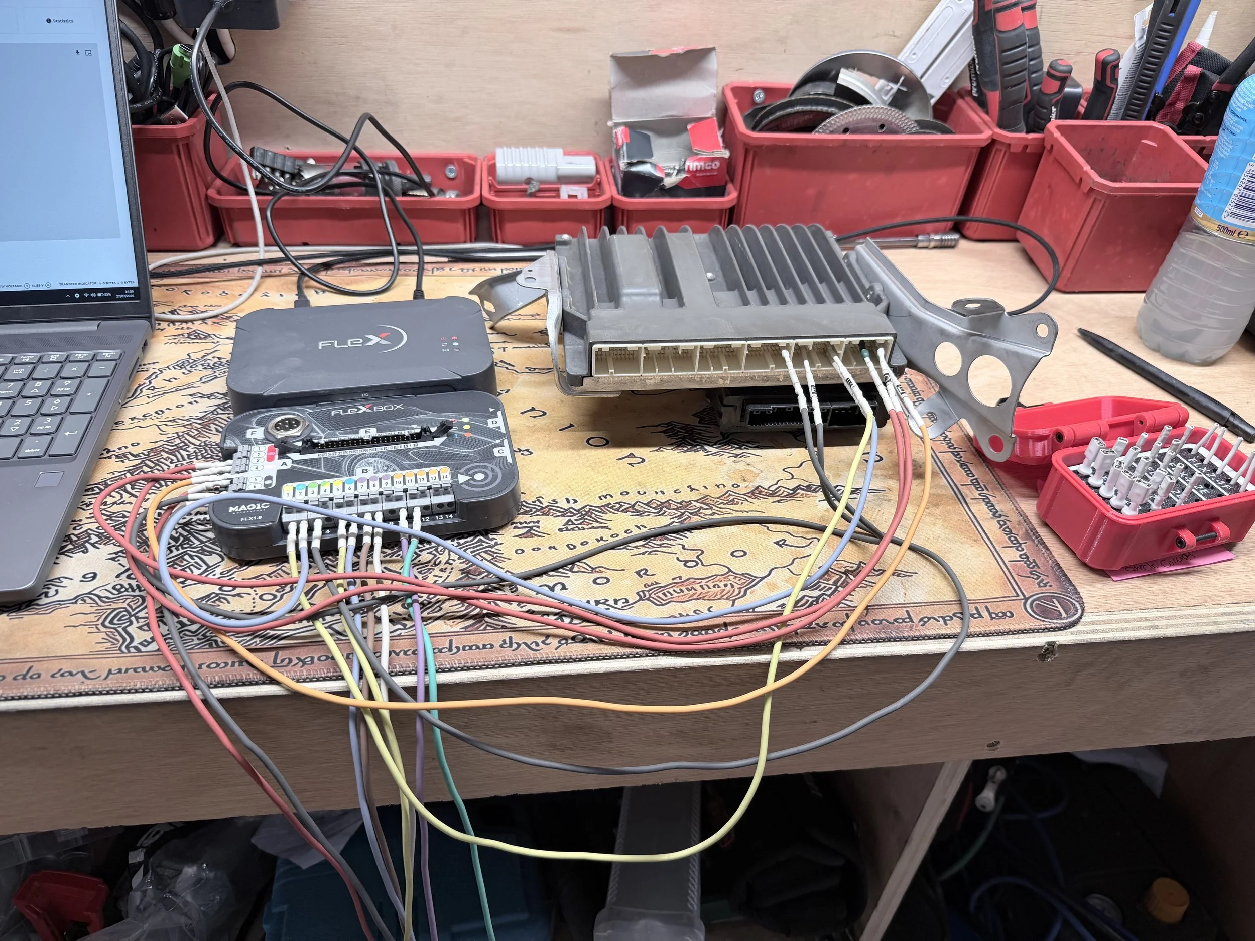 Workstation with electronic components, tools, and wiring including a laptop, an electronic control module, a wiring harness, and various tools in red trays, set on a wooden workbench. Tuned by Nightfall Automotive