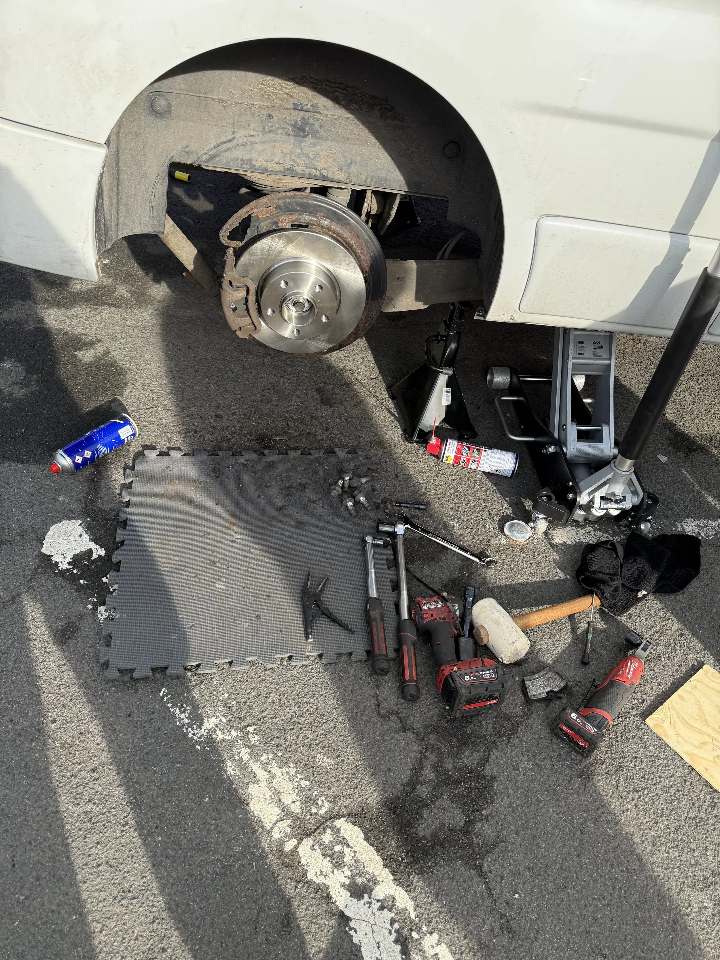 A car is lifted with a hydraulic jack, with its front wheel removed. Tools and parts, including a cordless drill, ratchets, sockets, and a tool mat, are laid out on the asphalt ground near the car, Breaks Fitted By Nightfall Automotive in Suffolk