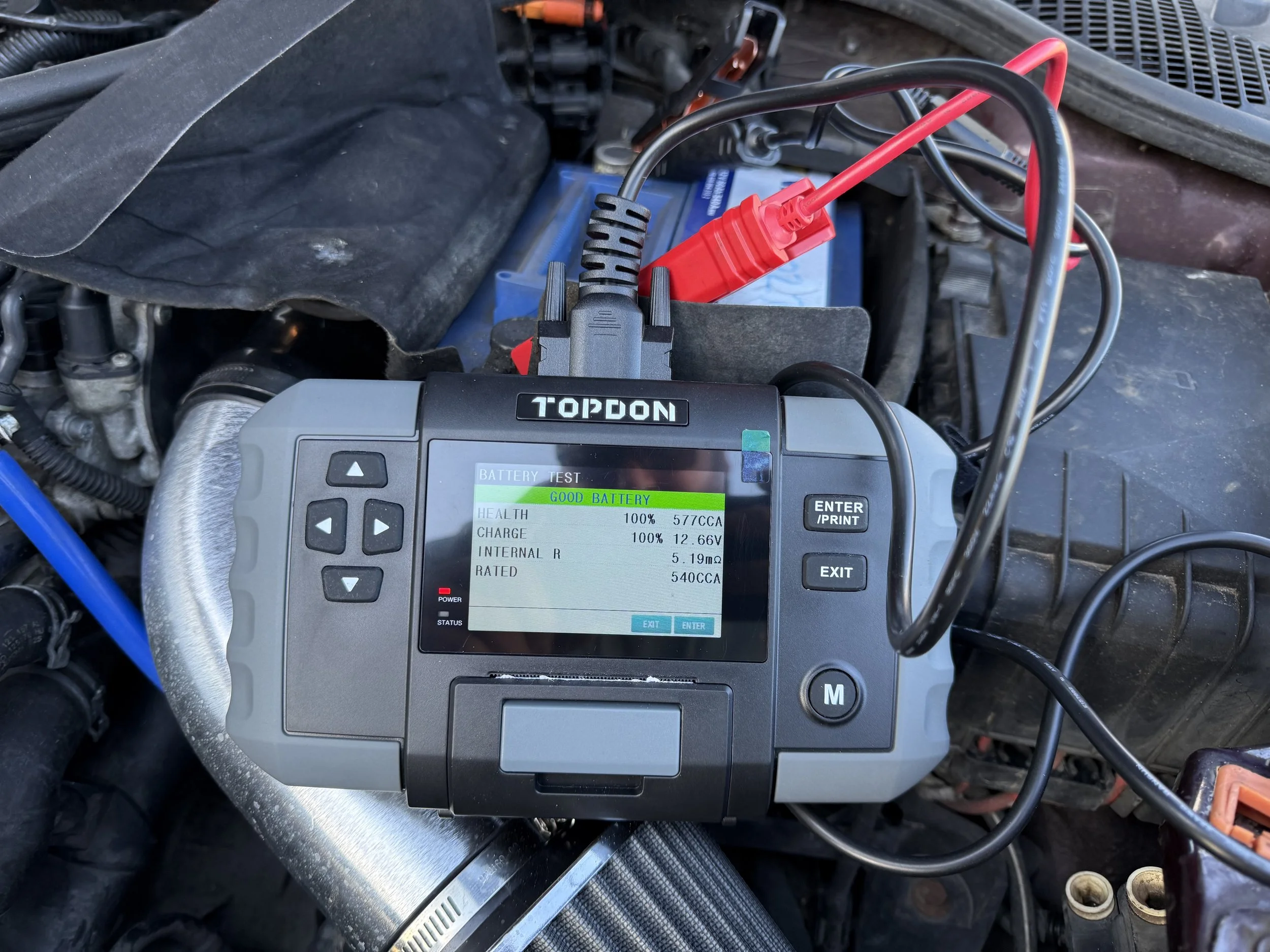 A battery tester connected to a car battery inside the engine bay, displaying battery health status as good with 100% charge and voltage of 12.66V.