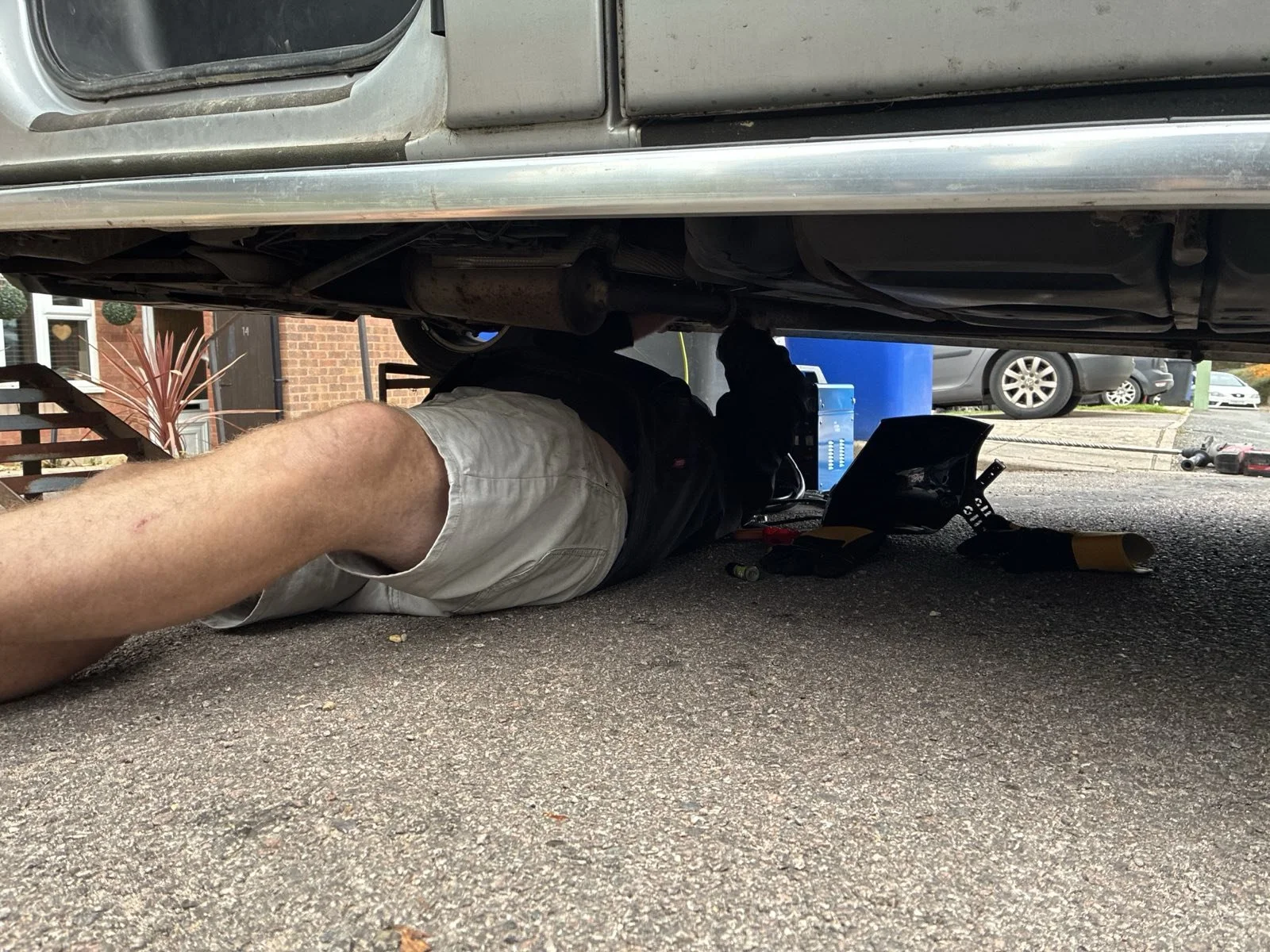 A person lying on their back working underneath a raised vehicle on a paved surface in a parking lot.