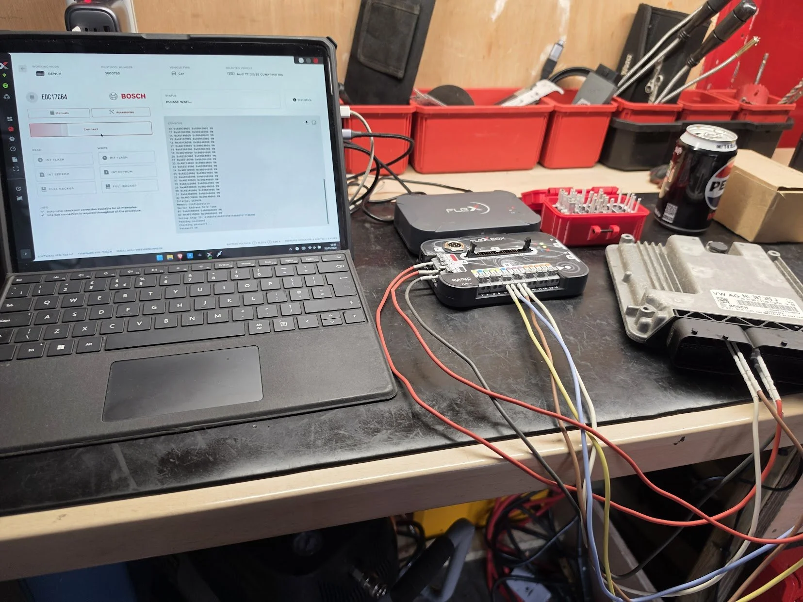 Laptop connected to automotive electronic repair tools and wires on a workbench, with tools and equipment in the background.