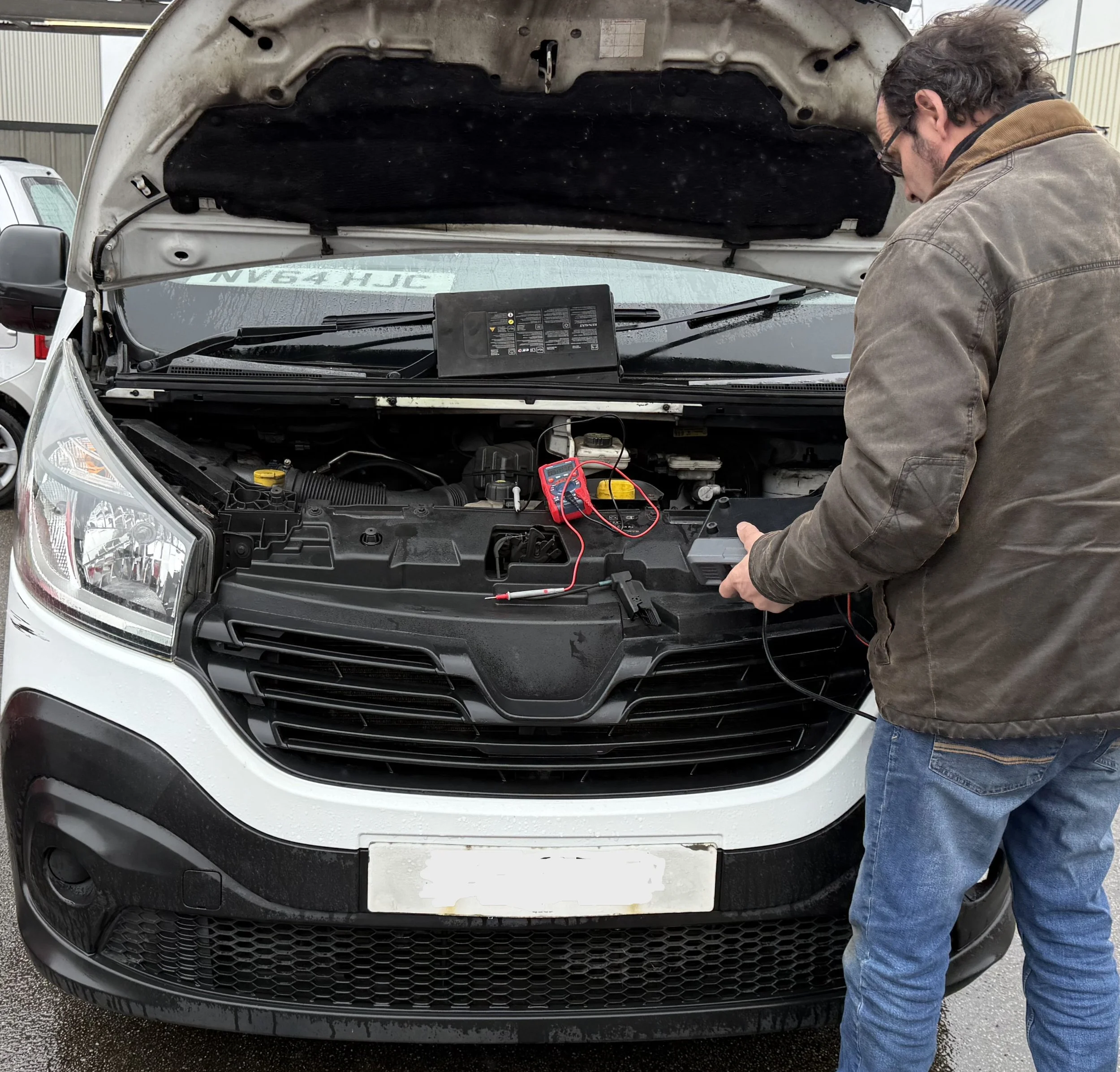 Man with dark hair and glasses working on the engine of a white van with the hood open, using electrical testing equipment in an outdoor parking lot on a rainy day.