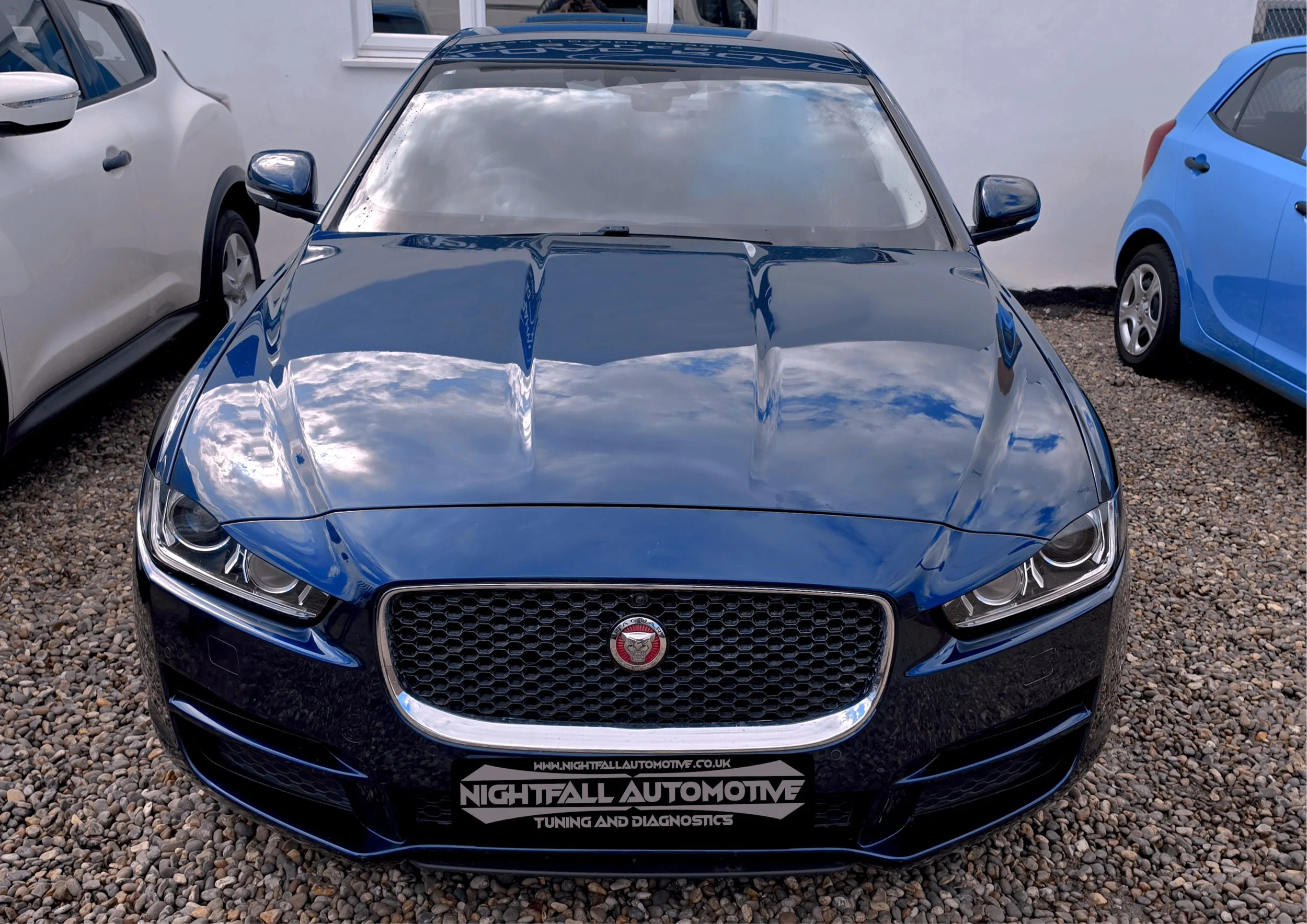 Front view of a dark blue Jaguar car parked on gravel, flanked by white and blue cars, with a dealership sign reading 'Nightfall Automotive' in the foreground. Tuned by Nightfall Automotive